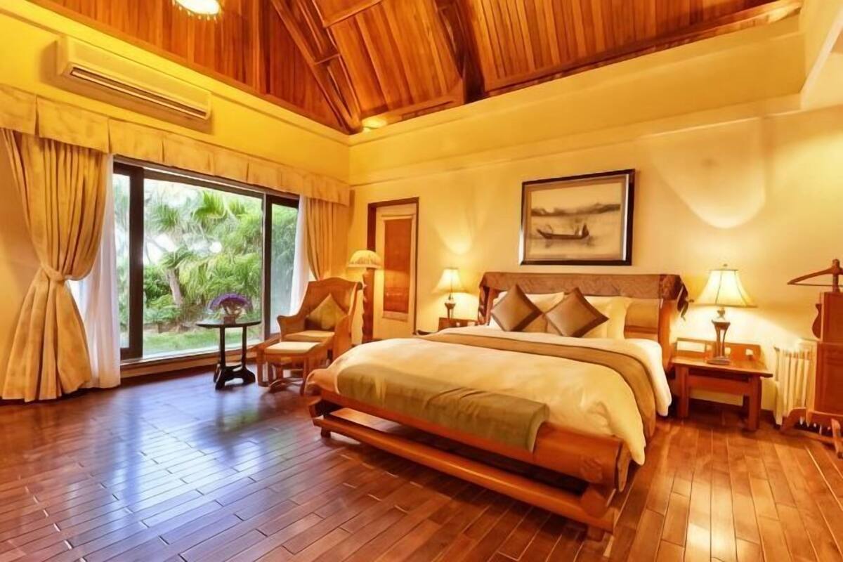 Un dormitorio amplio con una cama grande, suelo de madera, vistas por la ventana y un ventilador de techo.
