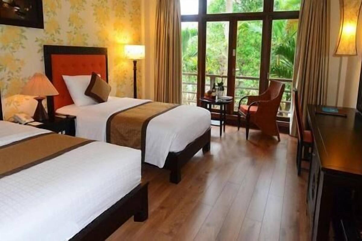 Habitación de hotel con dos camas, una mesita, una silla y vistas a zonas verdes a través de amplios ventanales.