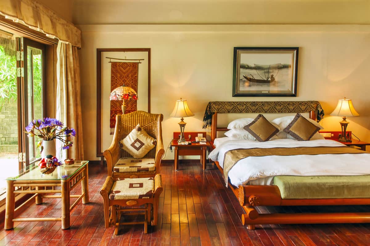 Una habitación de hotel con piso de madera, una cama con almohadas, una silla, una mesa y un ventanal con cortinas.