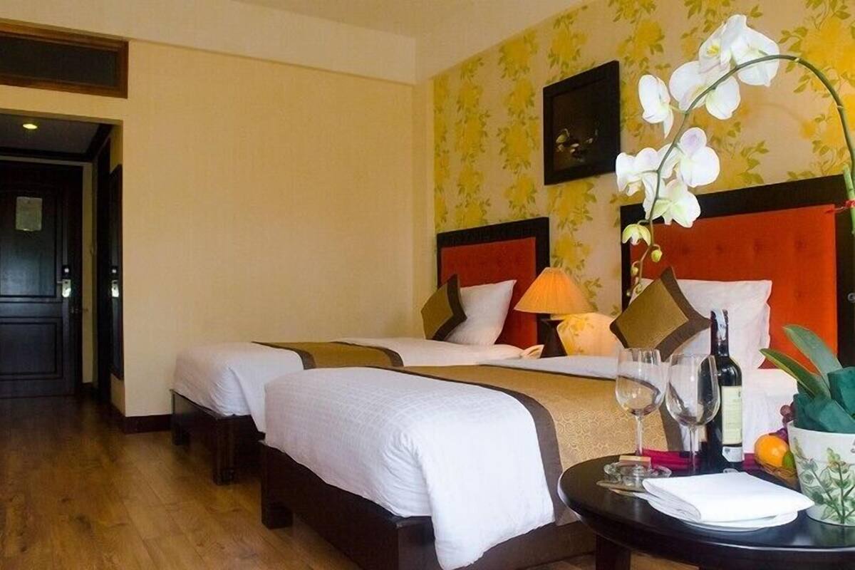 Una habitación de hotel con dos camas, una mesita con una botella y vasos, y un florero con flores.