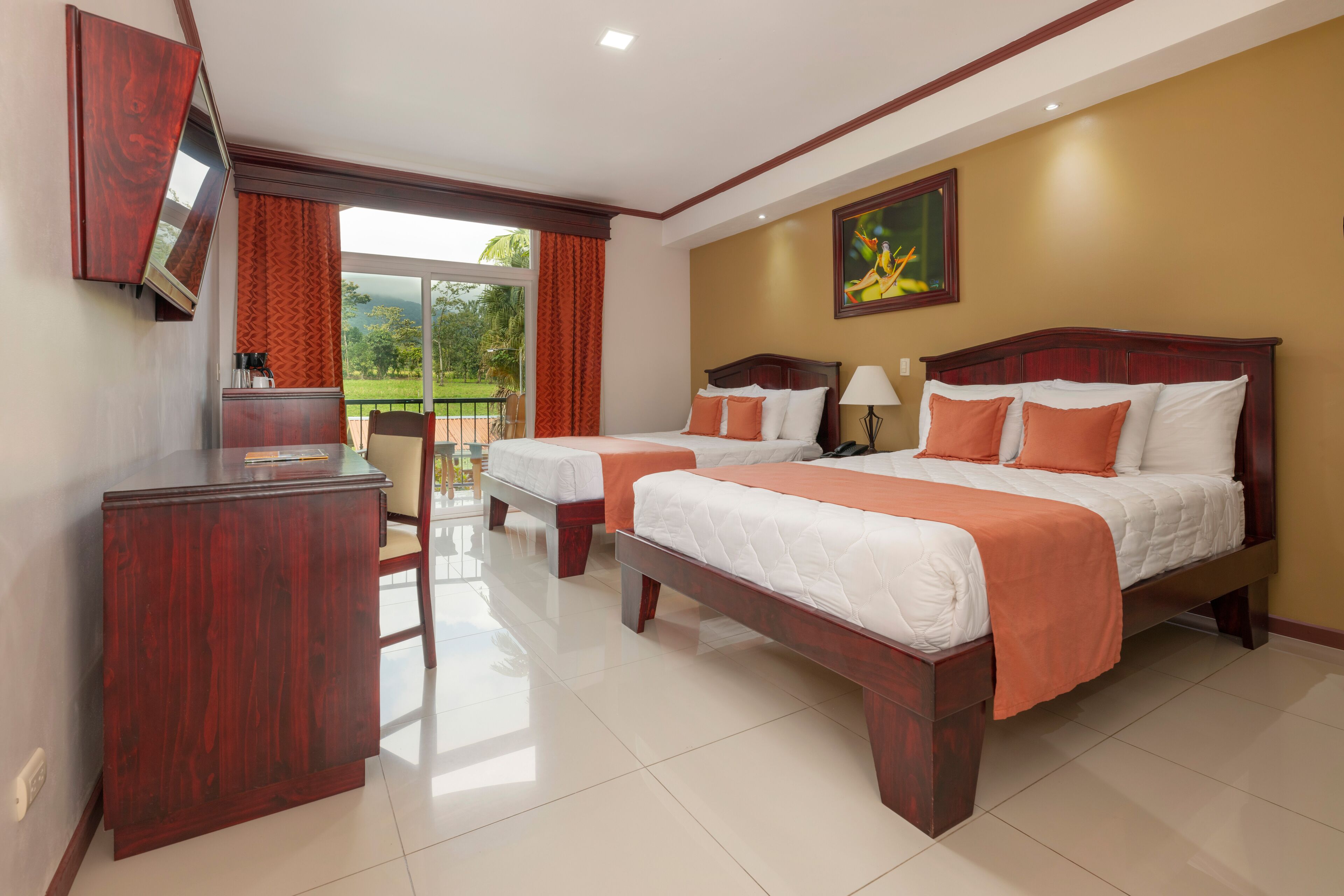 A hotel room with two beds, a TV, a desk, and a balcony with a view of the outdoors.