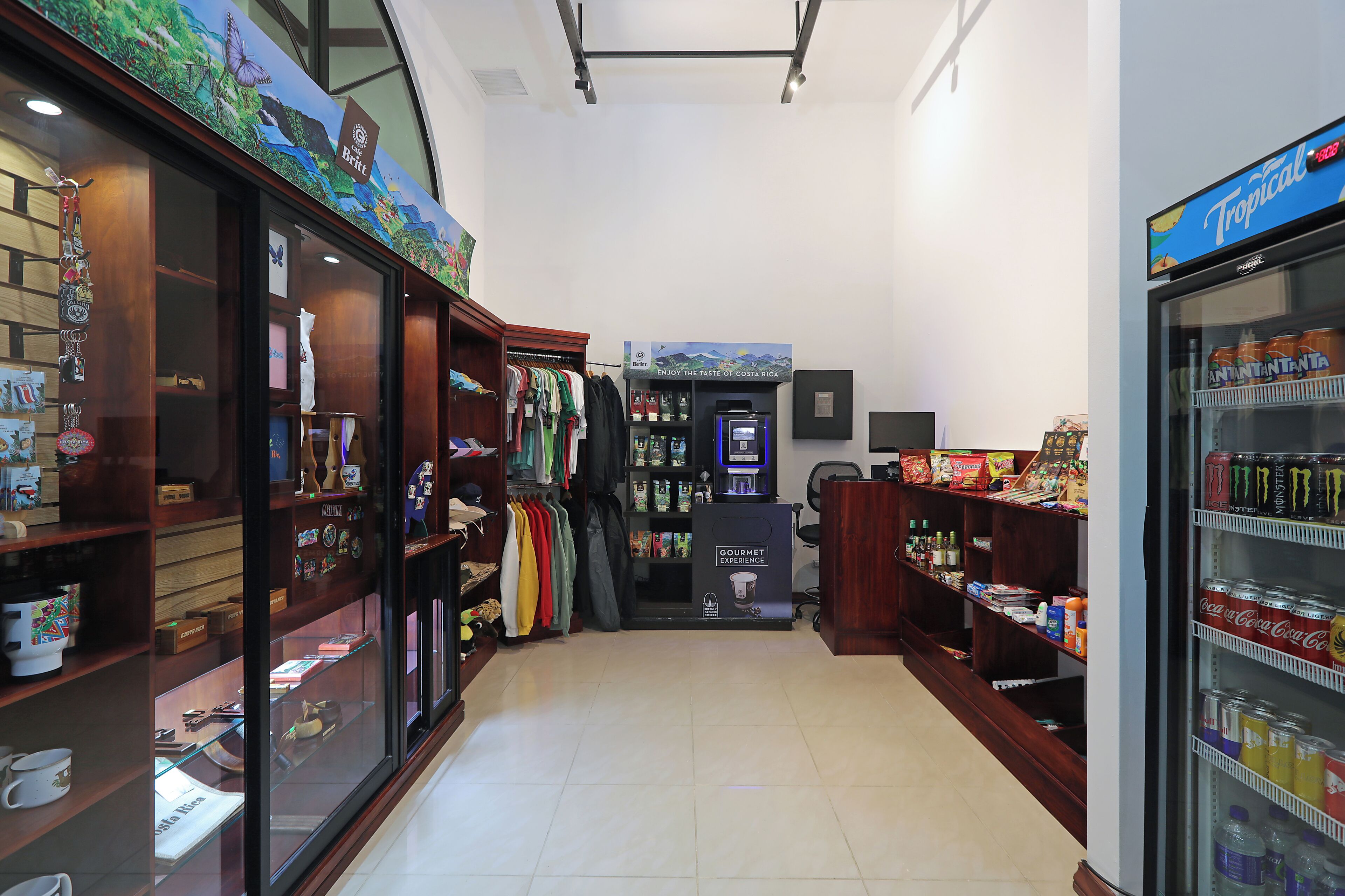 A store with shelves of snacks, a refrigerated drink display, and a coffee shop area with mugs and pastries.