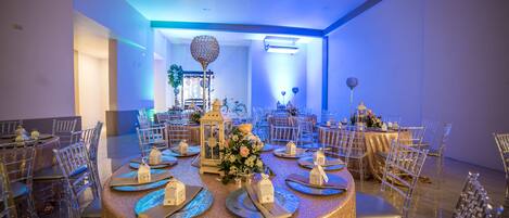 A banquet hall with round tables, clear chairs, and a chandelier.