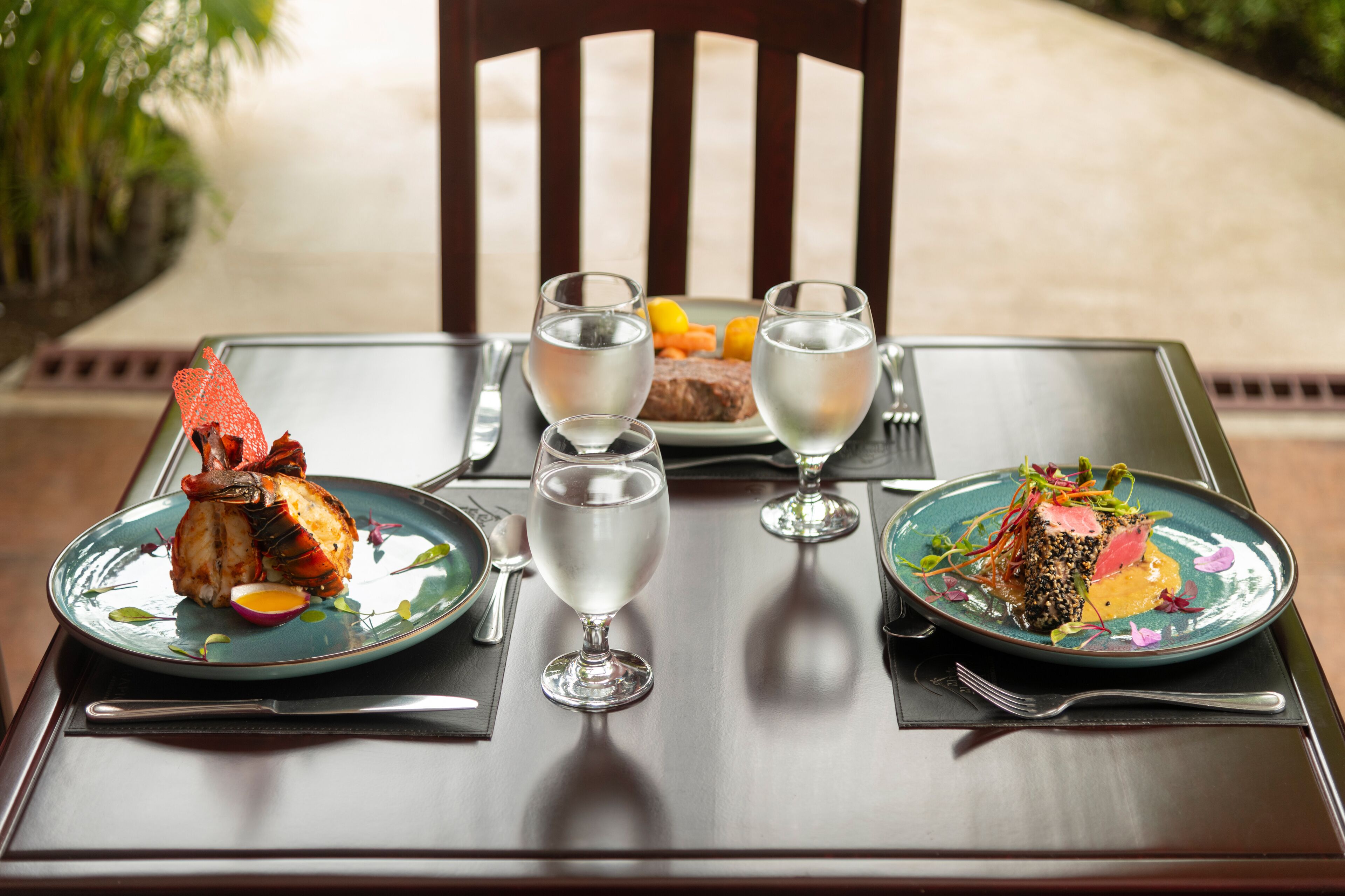 A dining table with two plates of food, two wine glasses, and a plate of fruit.
