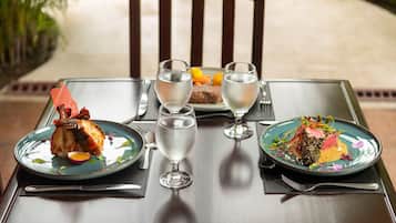 A dining table with two plates of food, two wine glasses, and a plate of fruit.