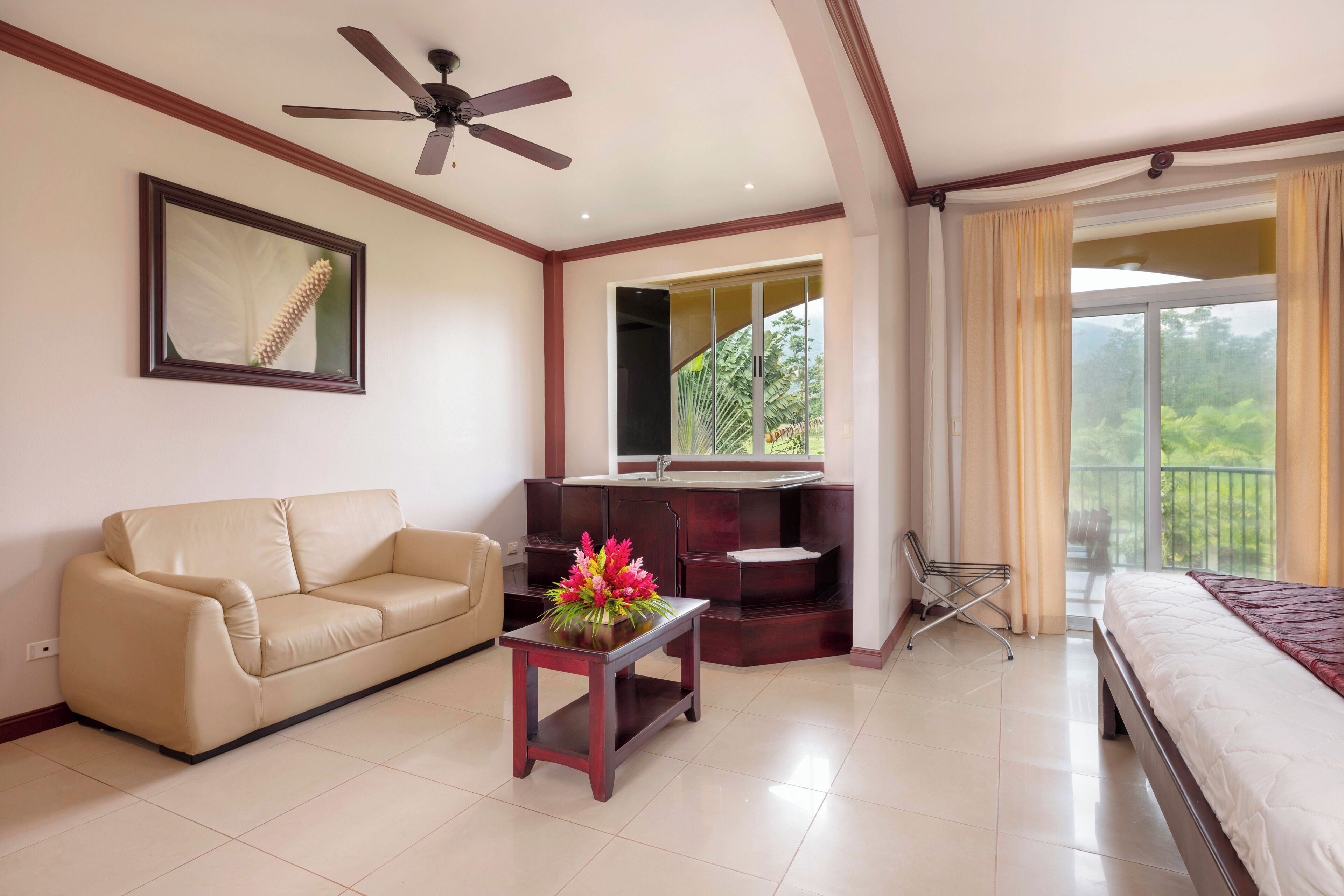 A hotel room with a sofa, a small table with flowers, a bed, a ceiling fan, a large window with curtains, and a bathroom with a sink and mirror.