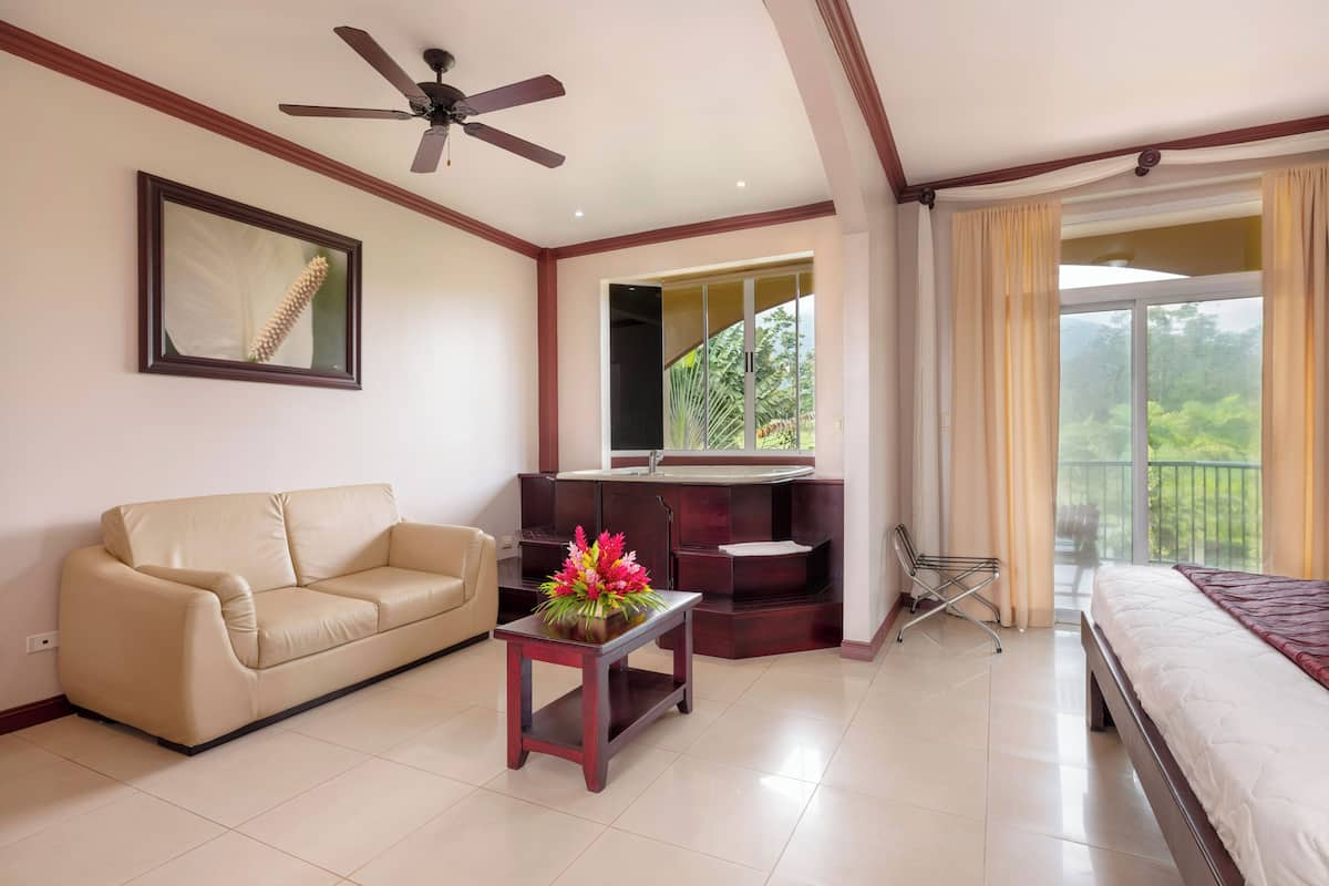 A hotel room with a sofa, a small table with flowers, a bed, a ceiling fan, a large window with curtains, and a bathroom with a sink and mirror.