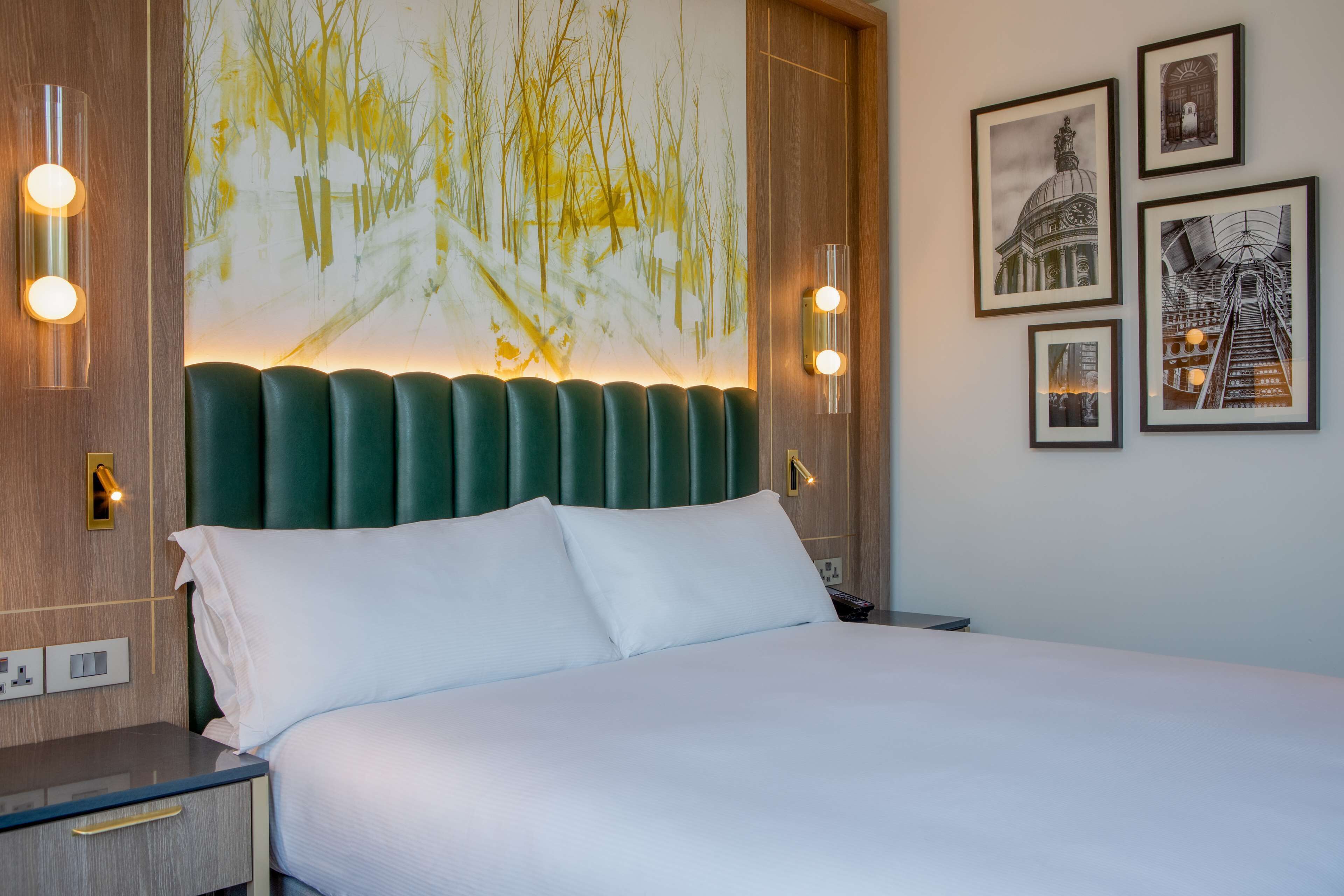 A hotel room with a large bed, a green upholstered headboard, and a wall-mounted artwork depicting a snowy landscape.