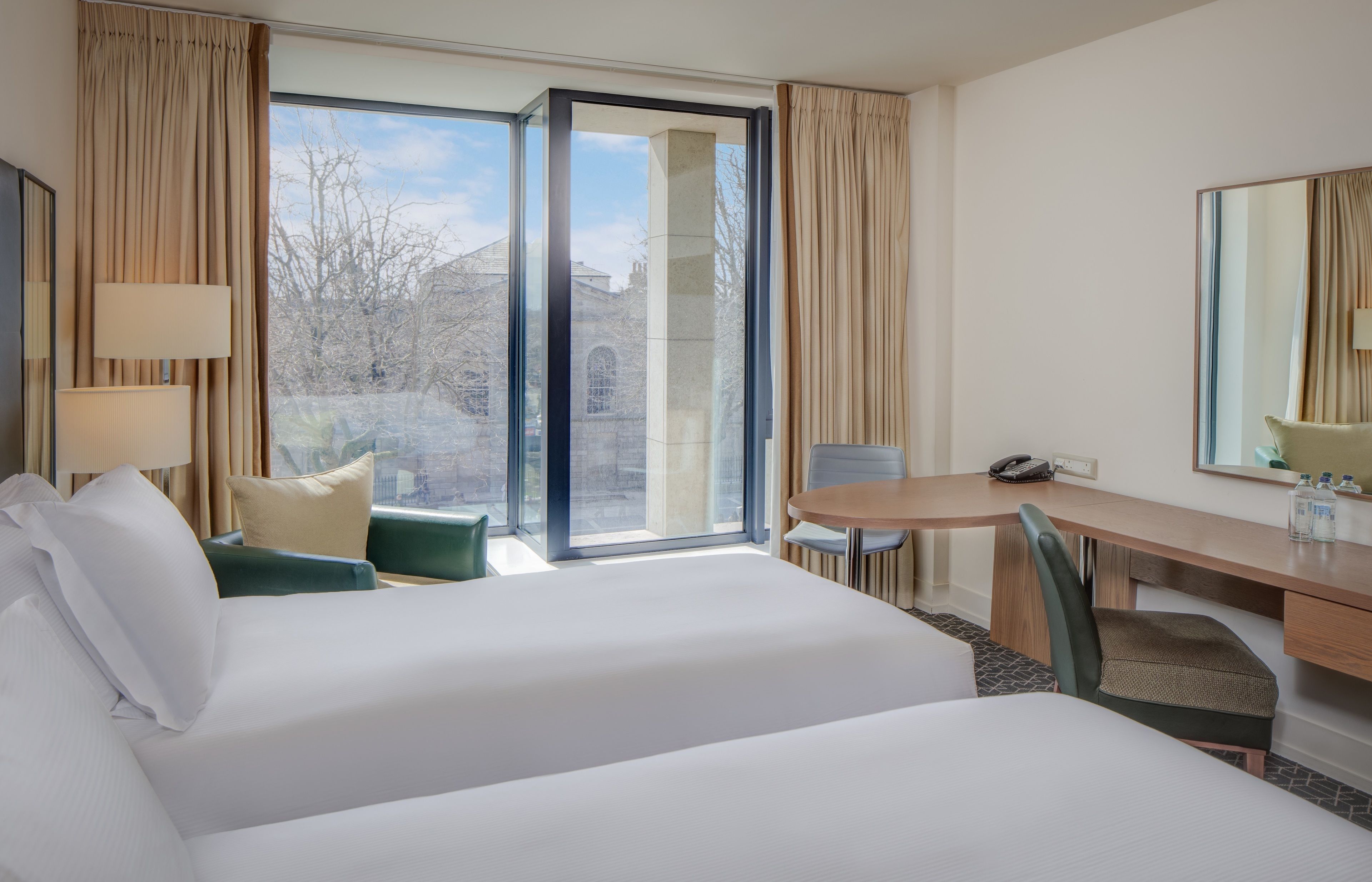 A hotel room with a large bed, a desk, a chair, and a view of a building through a glass door.