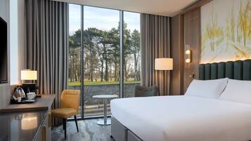 A hotel room with a large bed, a desk with a chair, a small table, and a view of a park through large windows.