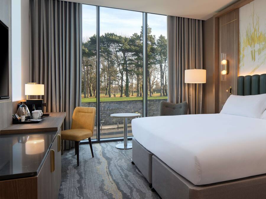 A hotel room with a large bed, a desk with a chair, a small table, and a view of a park through large windows.