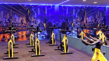 Une salle de sport avec des vélos d’appartement disposés en rangées, un mur recouvert de miroirs et un plafond éclairé d’une lumière bleue.