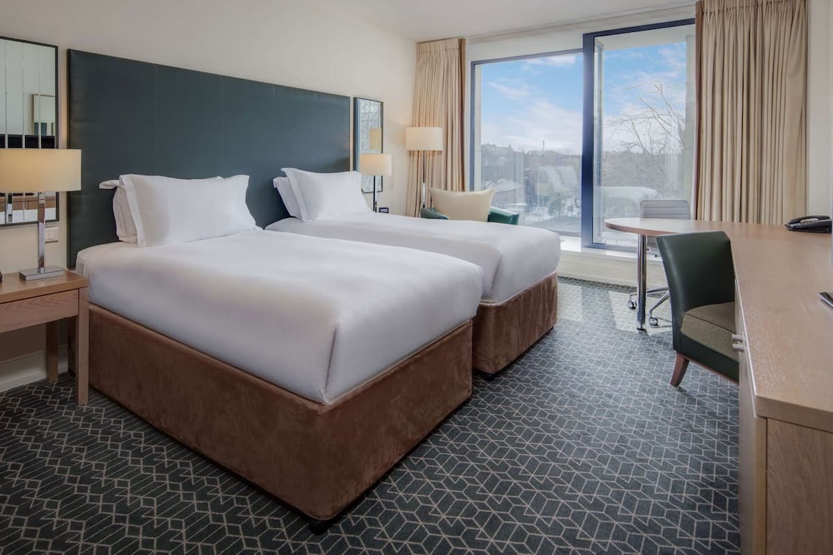 A hotel room with two beds, a desk with a phone, a chair, a TV, and a window with curtains.