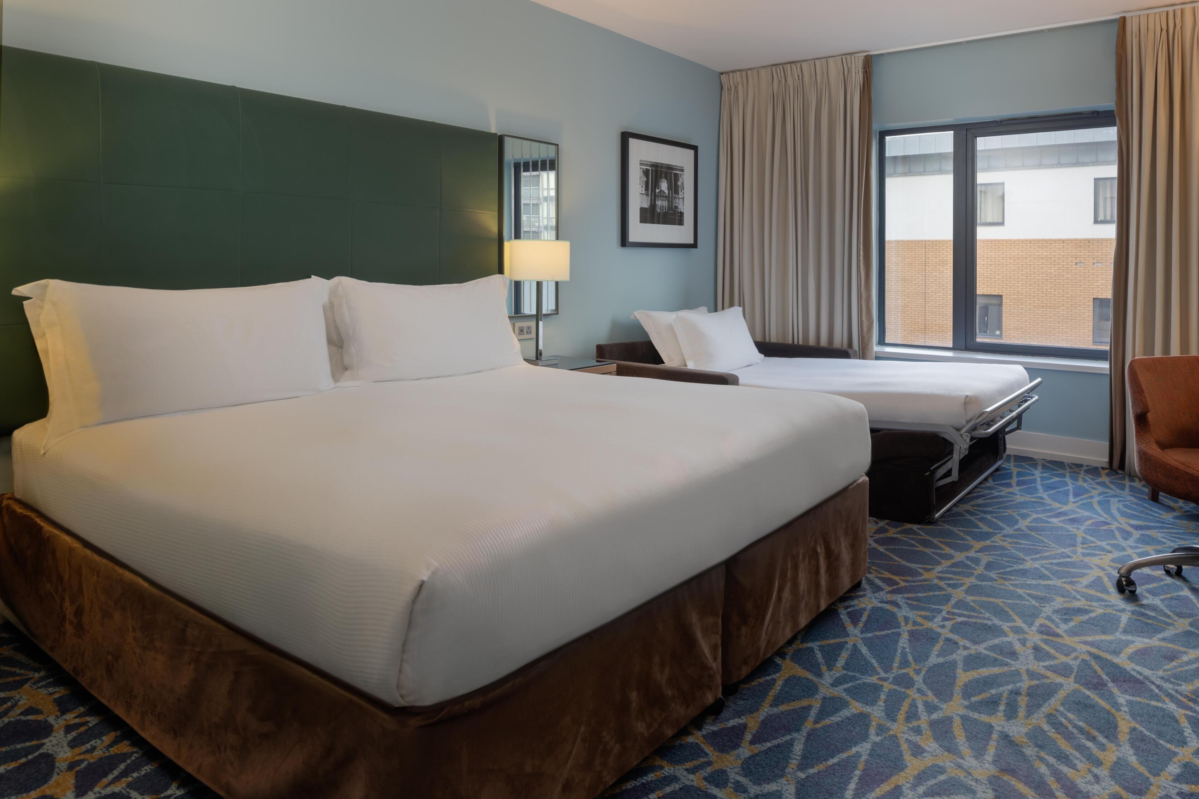 A hotel room with a large bed, a smaller bed, a desk, a chair, a lamp, and a window with curtains.