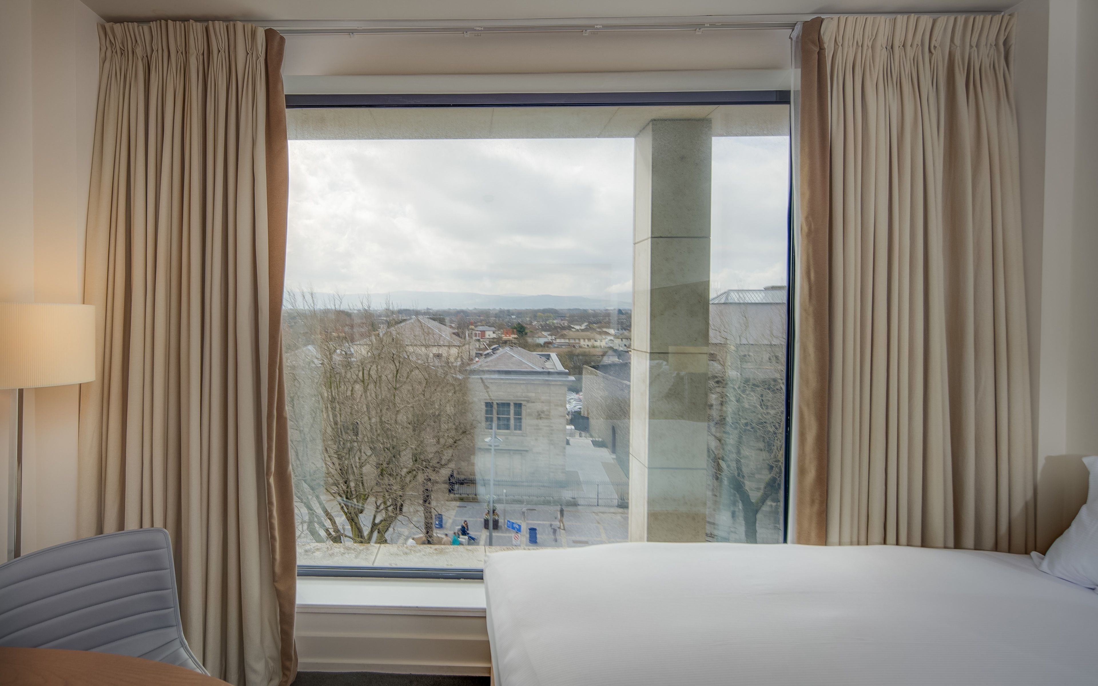 Una habitación de hotel con un ventanal grande, una cama, una silla y una lámpara.