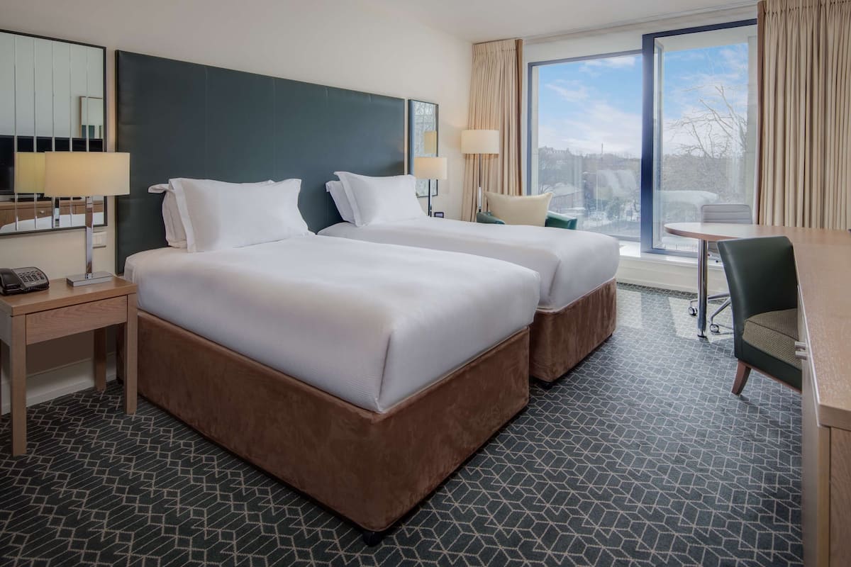 A hotel room with two beds, a desk, a chair, a TV, and a window with curtains.