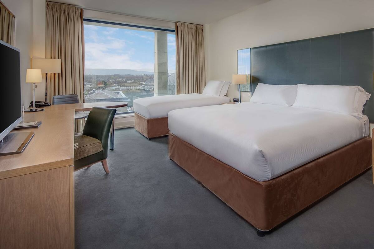 A hotel room with two beds, a desk with a computer, a chair, and a large window offering a city view.