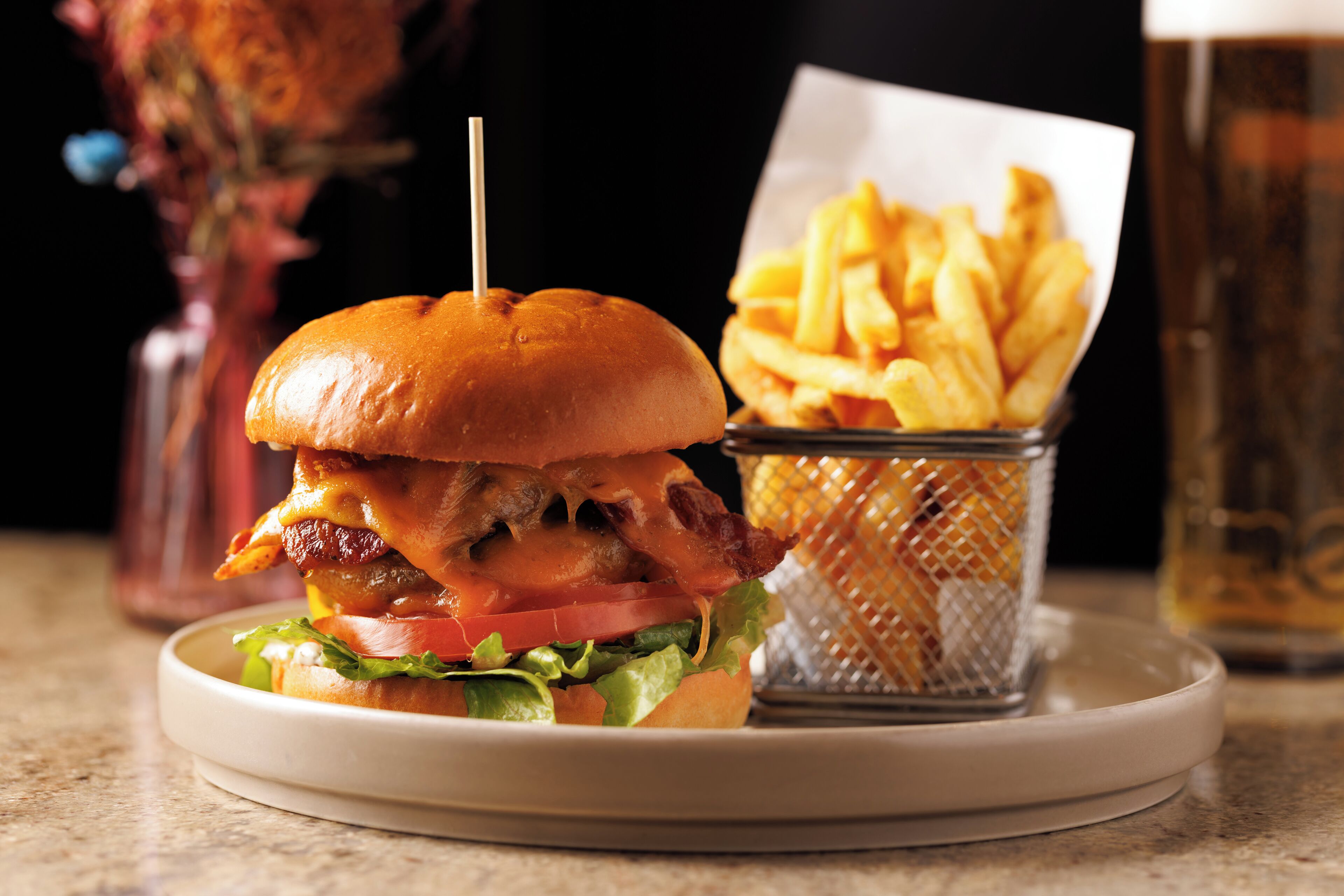 A burger with bacon, cheese, and lettuce on a bun, served with fries and a drink.