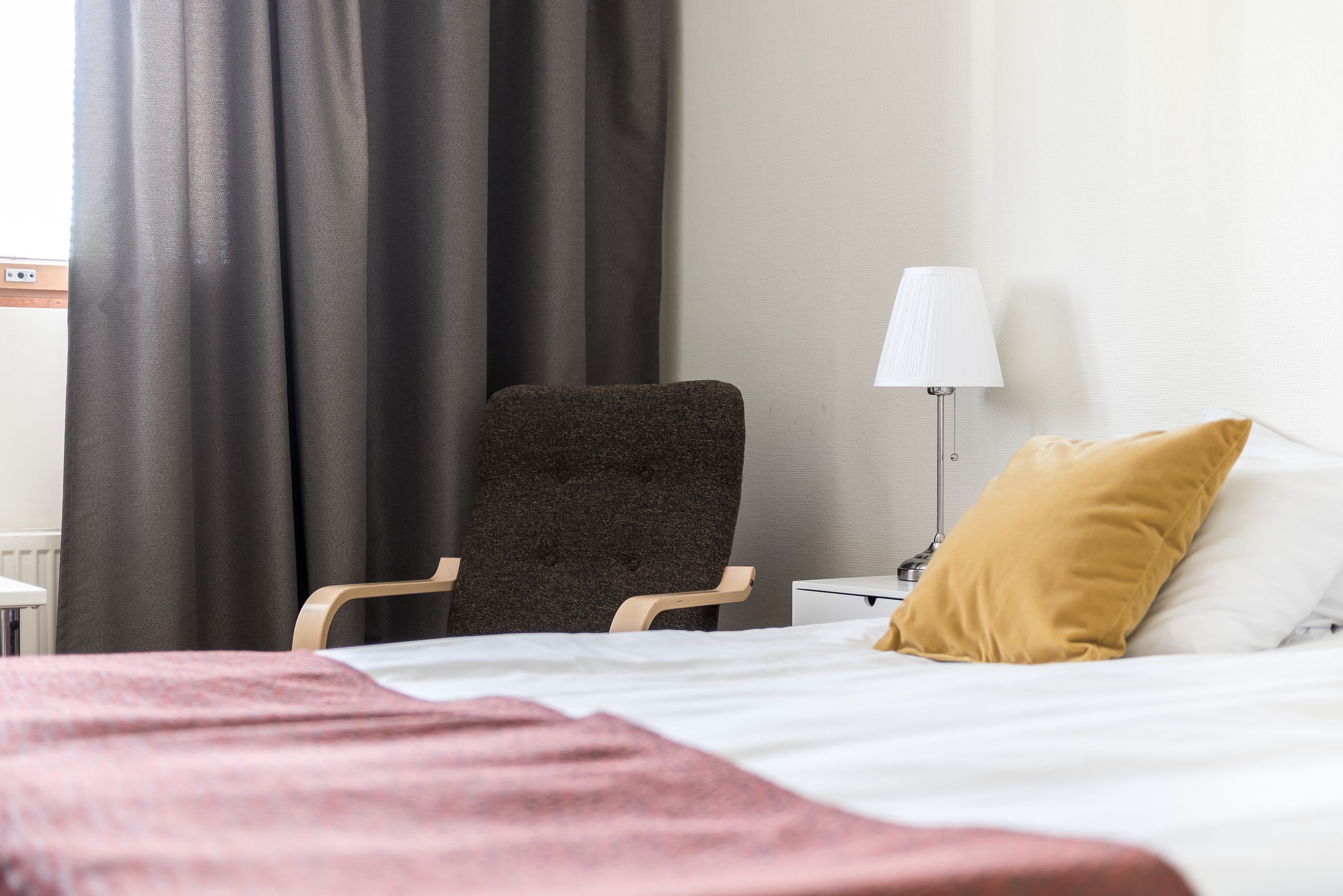 A hotel room with a bed, a chair, a lamp, and a window with curtains.