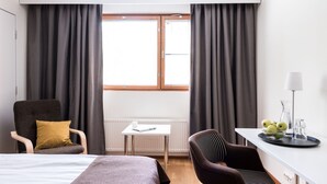 A hotel room with a bed, a desk, a chair, a radiator, and a window with curtains. - Hotel Hermica (Tampere)