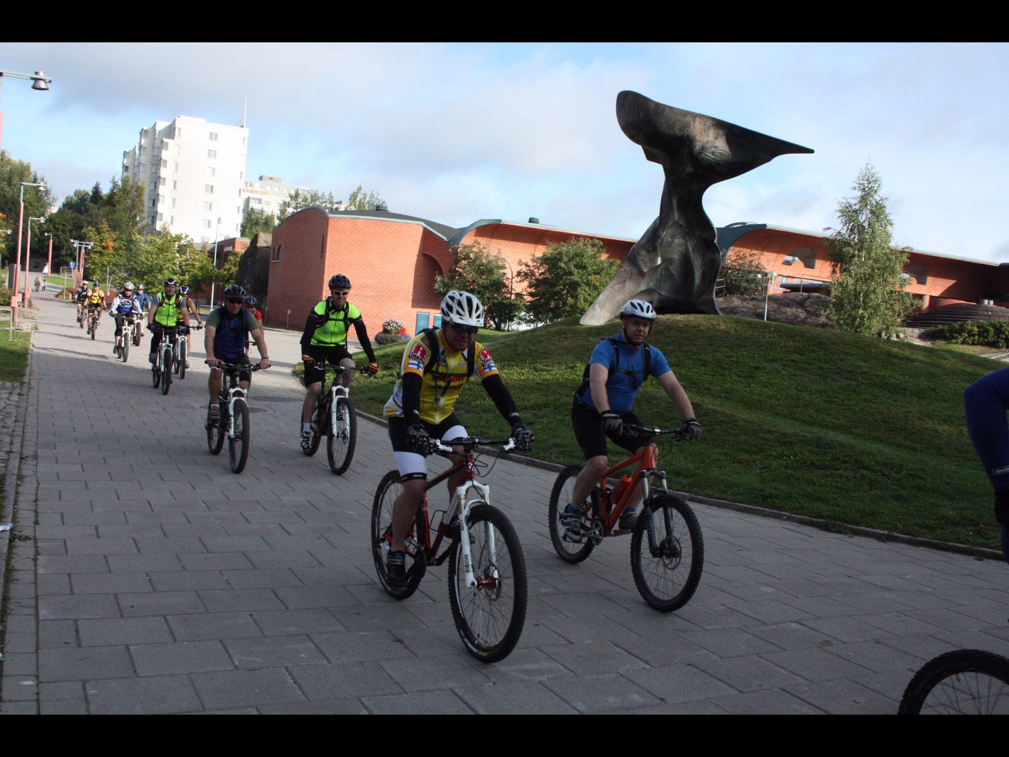 A group of cyclists on a paved path with a large sculpture in the background.