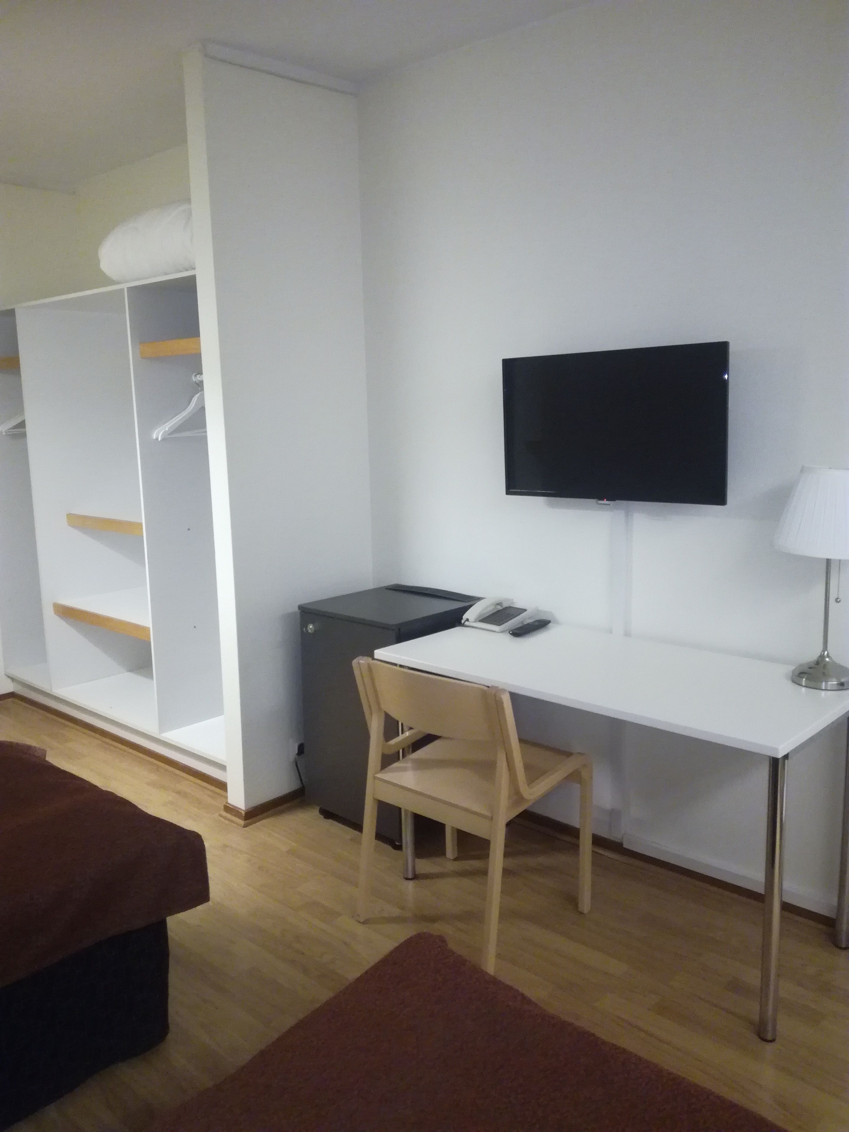A room with a white desk, a chair, a television mounted on the wall, a small cabinet, and a brown couch.