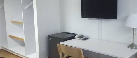 A room with a white desk, a chair, a television mounted on the wall, a small cabinet, and a brown couch.
