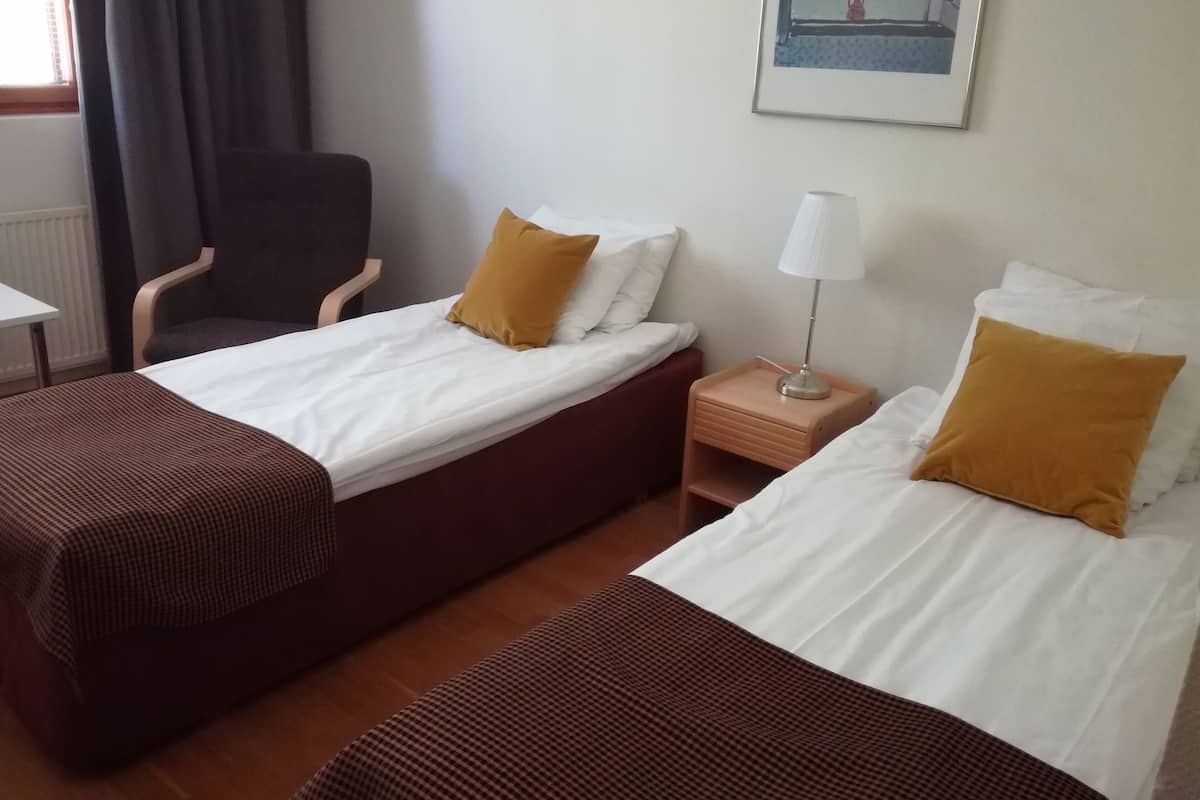 A hotel room with two single beds, a chair, a nightstand with a lamp, and a painting on the wall.