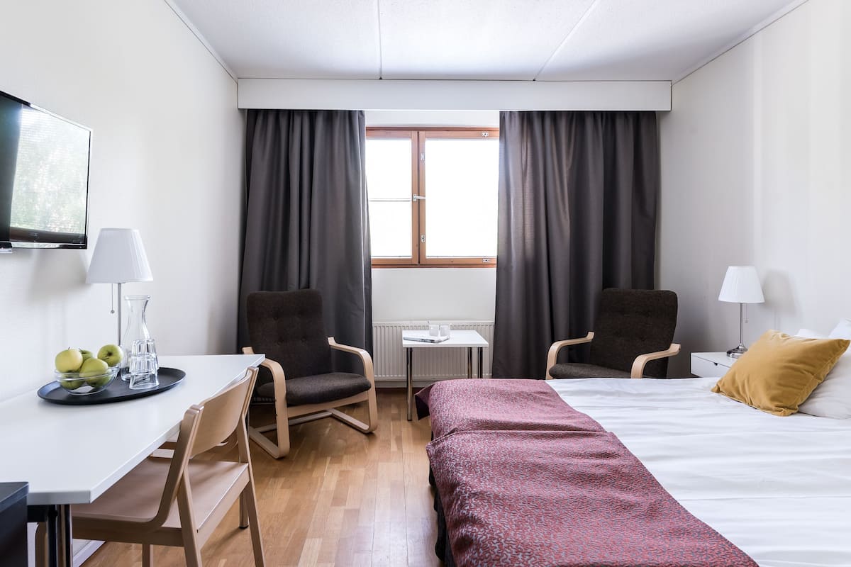A hotel room with a bed, two armchairs, a desk with a lamp, a TV, and a window with curtains.