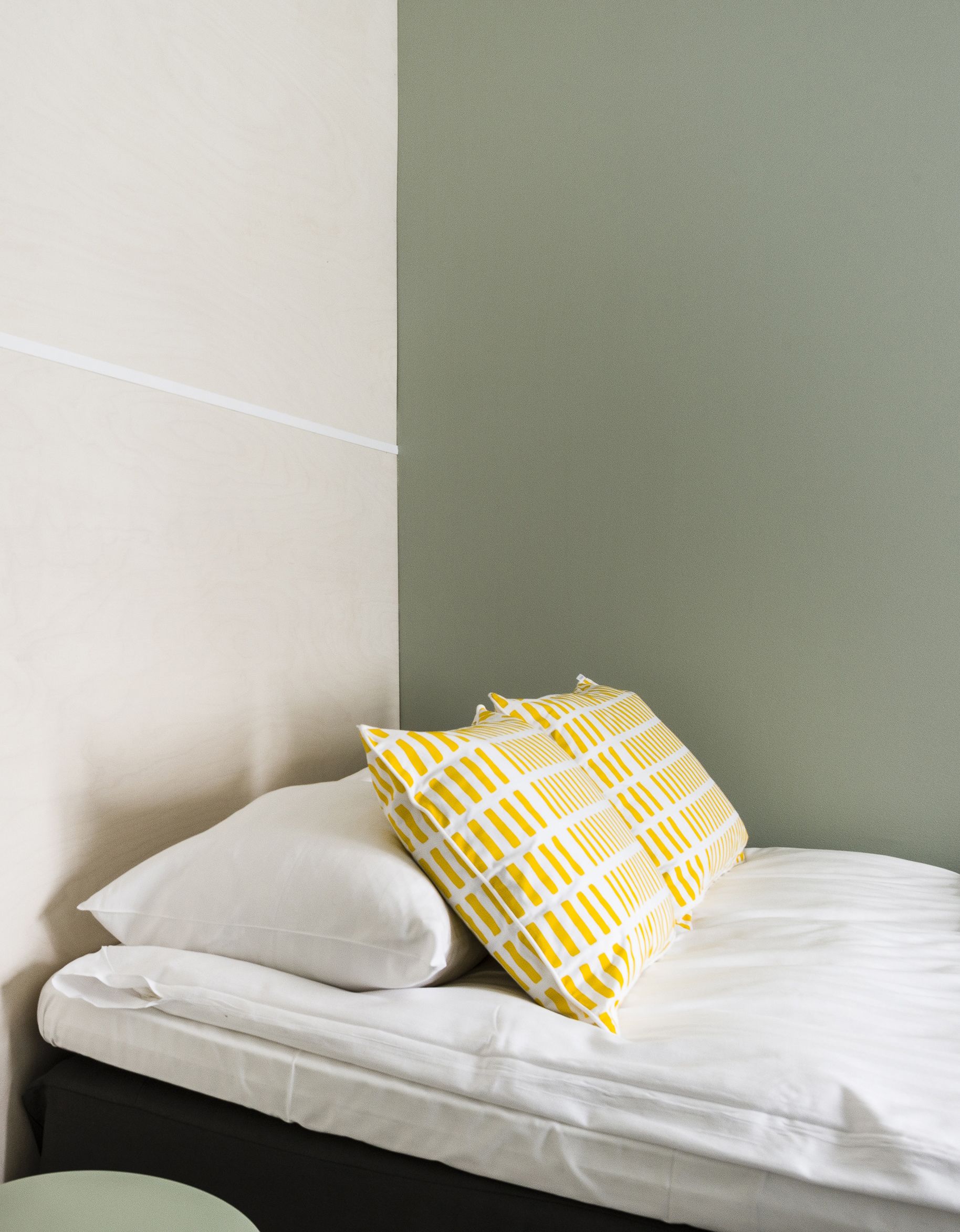 Una cama con ropa blanca y un cojín a rayas amarillo y blanco, frente a una pared de tonos verde claro y oscuro.