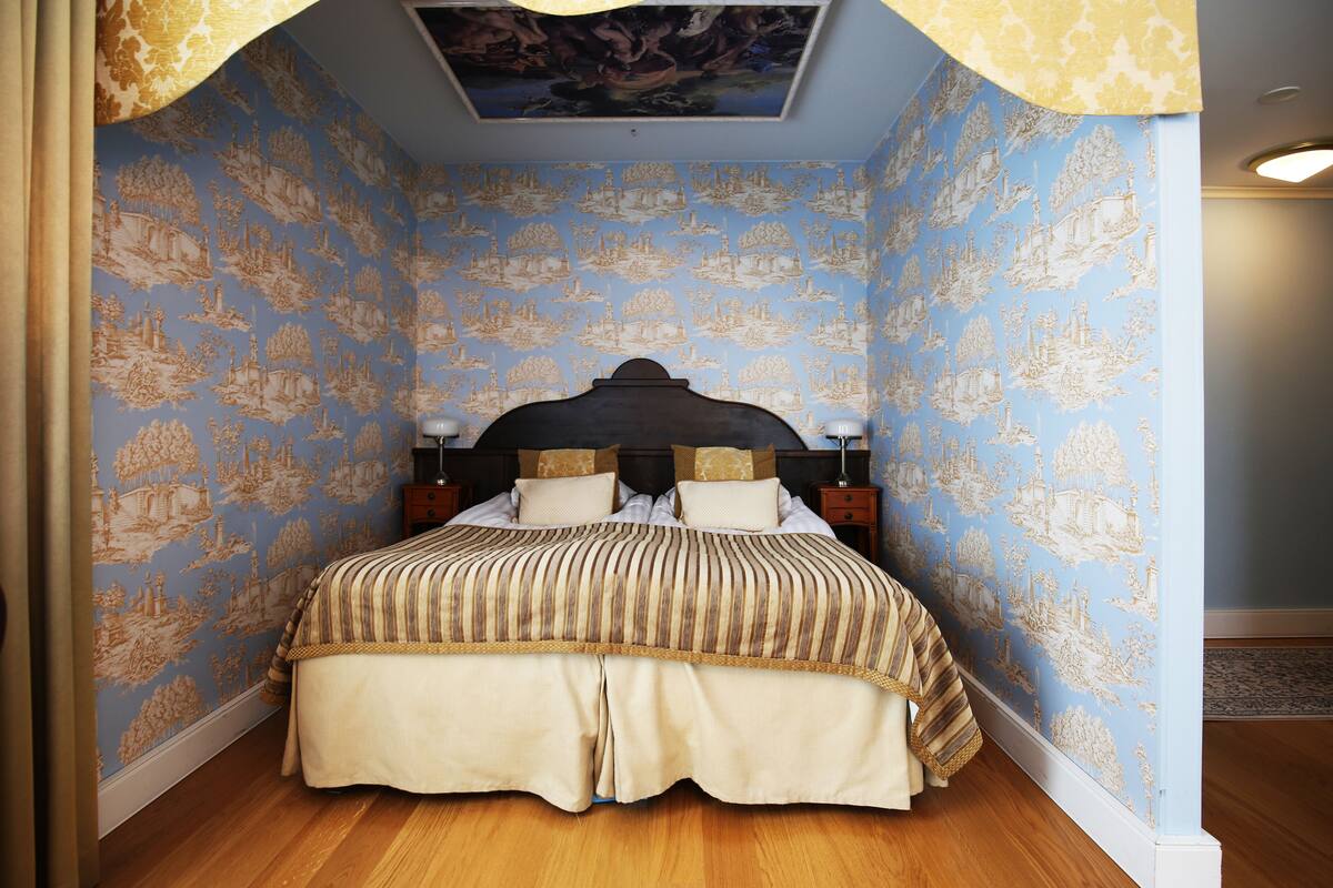 A bedroom with a large bed, patterned wallpaper, and a ceiling mural.