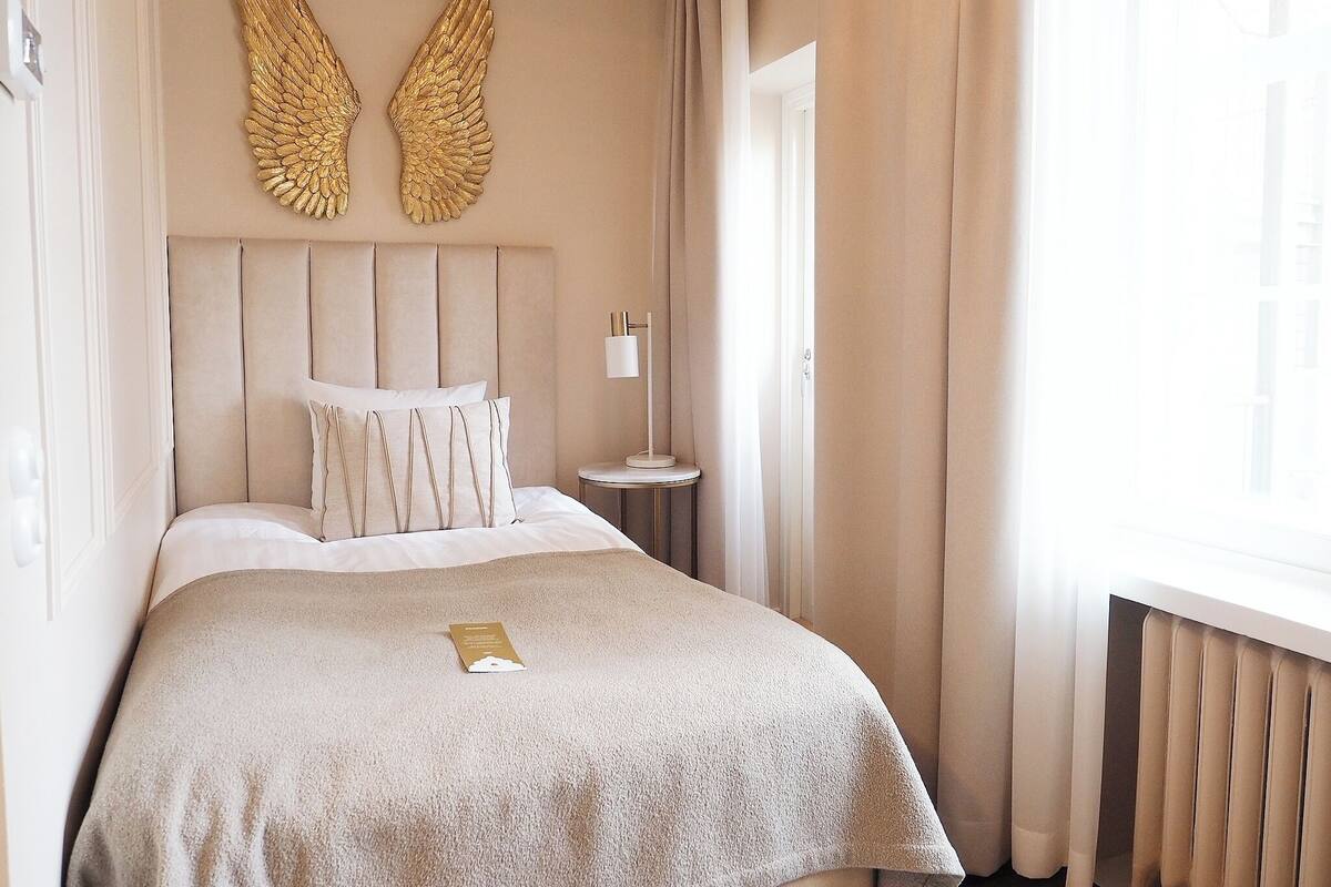 A hotel room with a large bed, a headboard with wing-shaped decorations, a bedside table with a lamp, and a window with curtains.