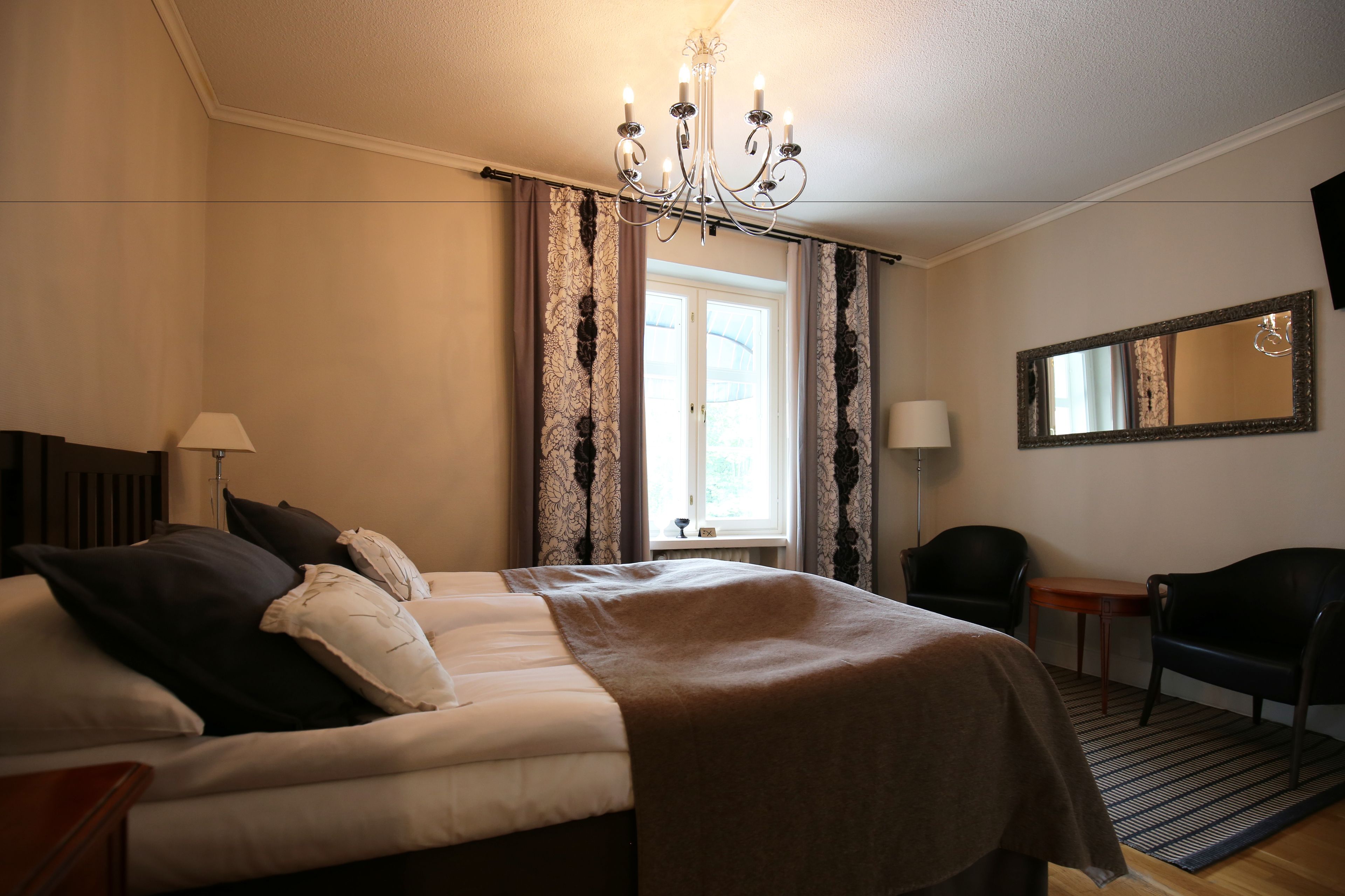 A hotel room with a bed, a chair, a small table, a lamp, a mirror, and a window with curtains.