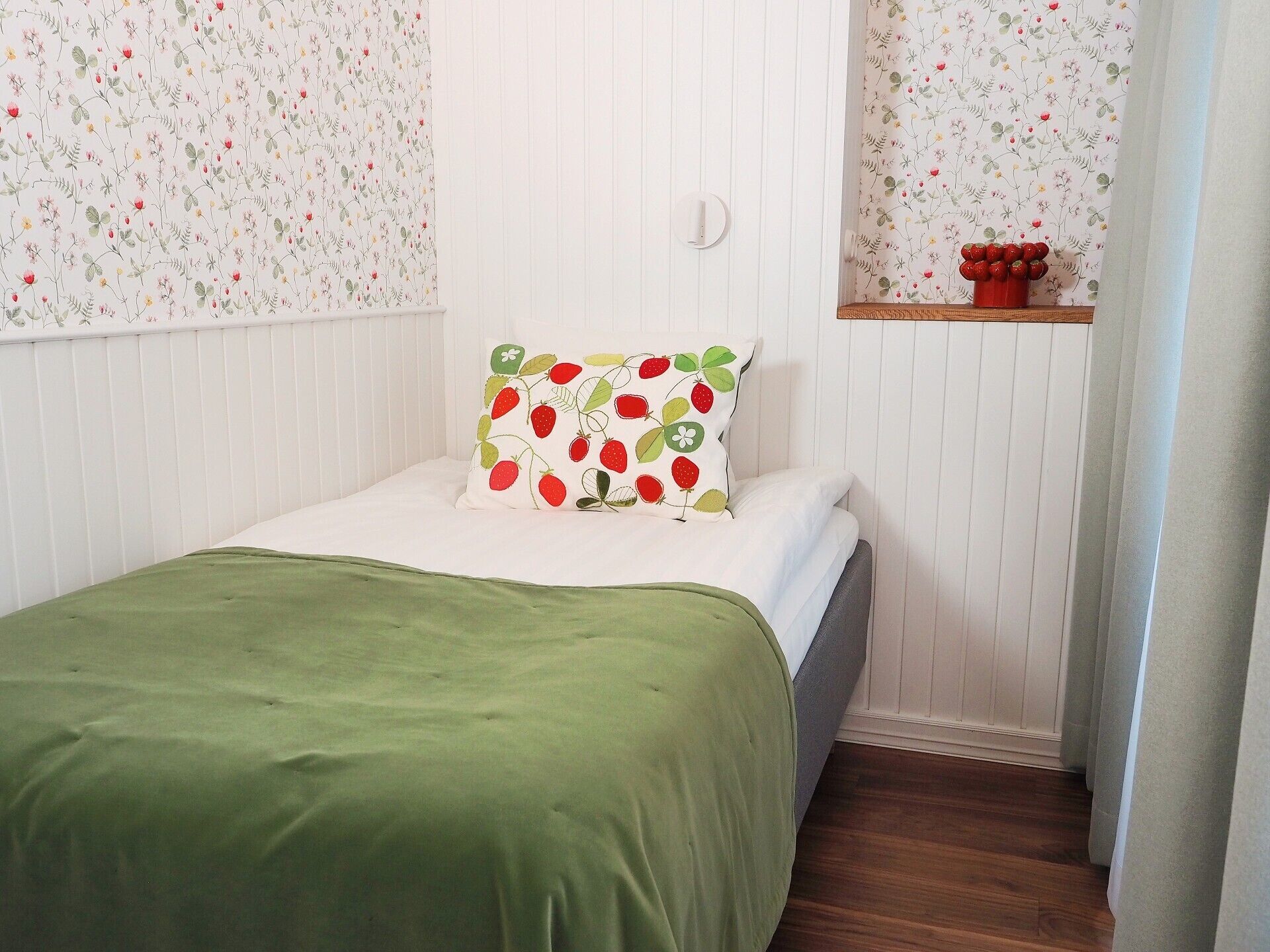 A small, neatly arranged bedroom with a single bed, a green bedspread, a floral-patterned pillow, and a small wooden shelf with a vase of red flowers.
