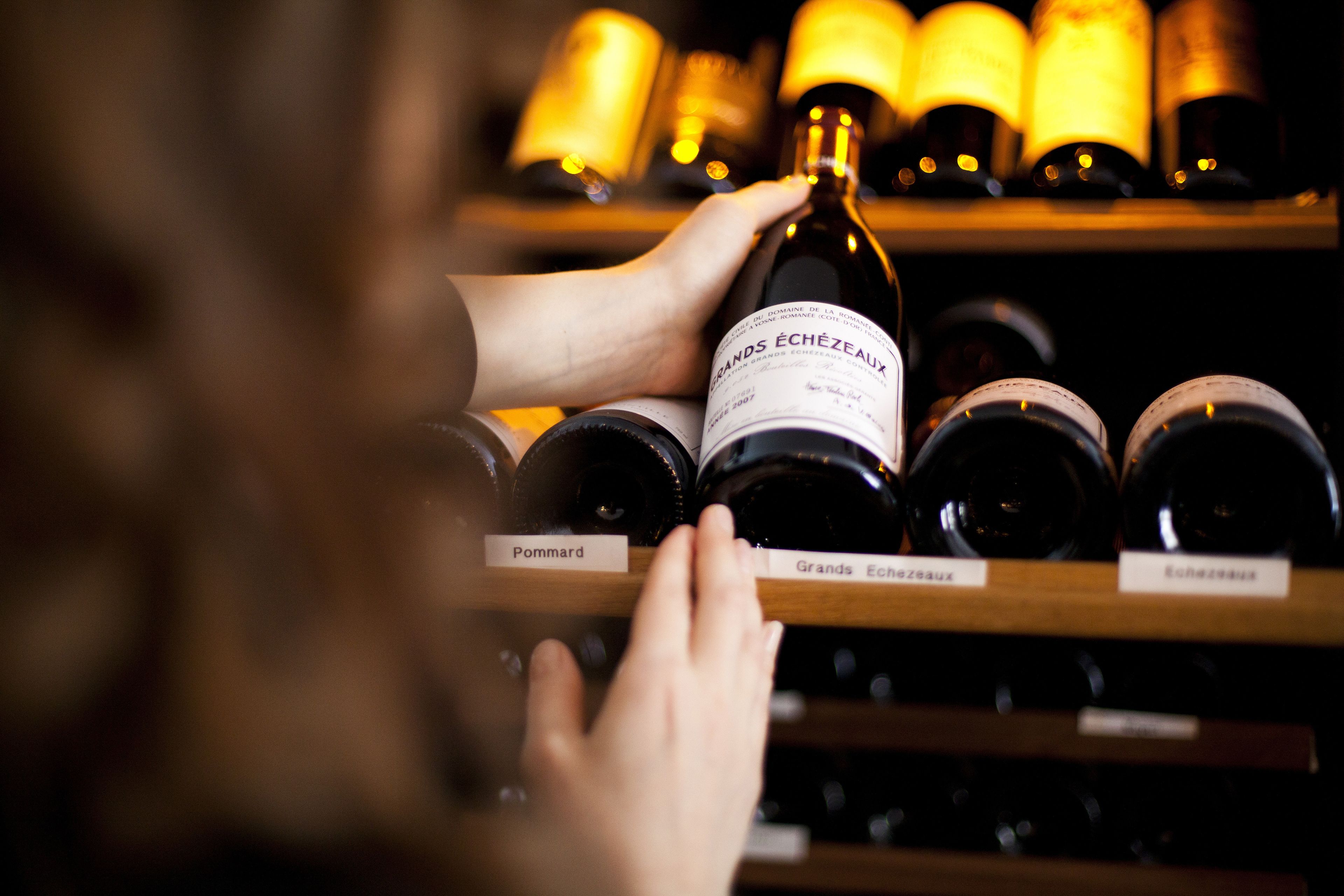 Una mano elige una botella de vino de un estante con varios nombres de vinos.