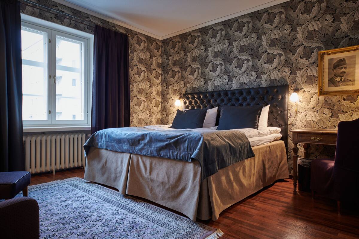A bedroom with a large bed, a radiator, a window with curtains, and a patterned wallpaper.