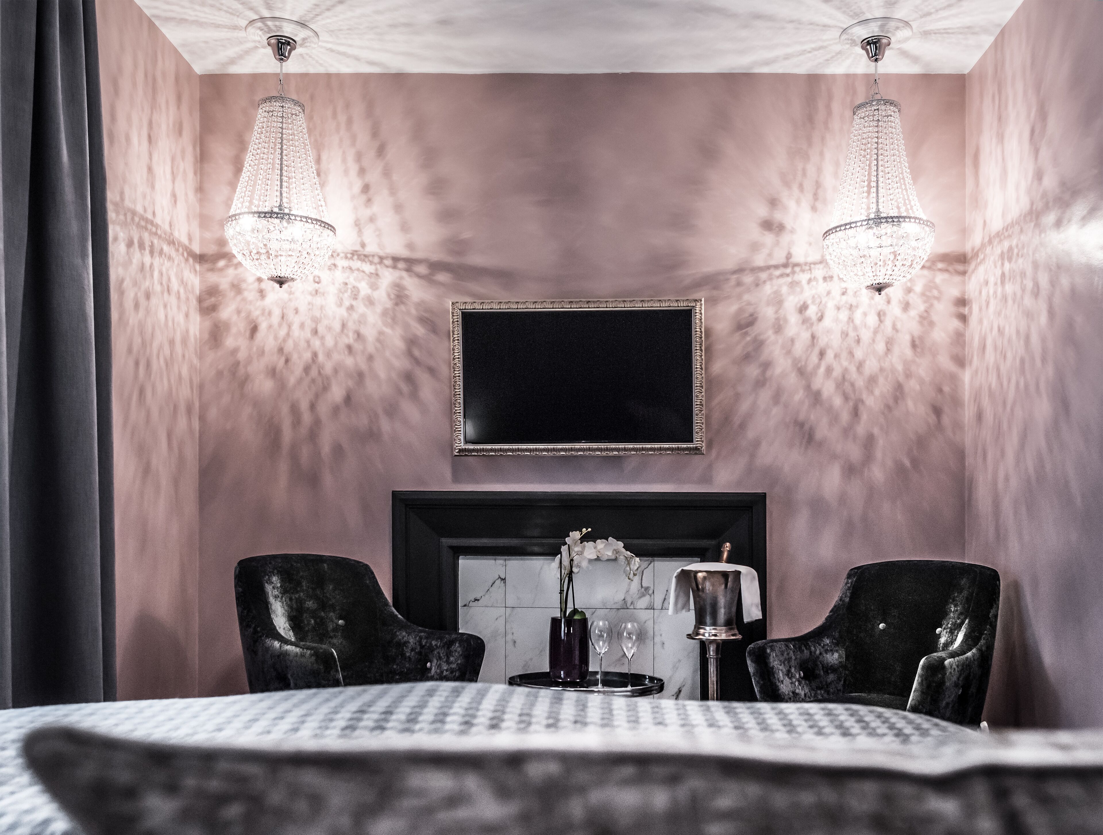 Une chambre à coucher avec un grand lit, deux fauteuils noirs, une cheminée et deux lustres en cristal.