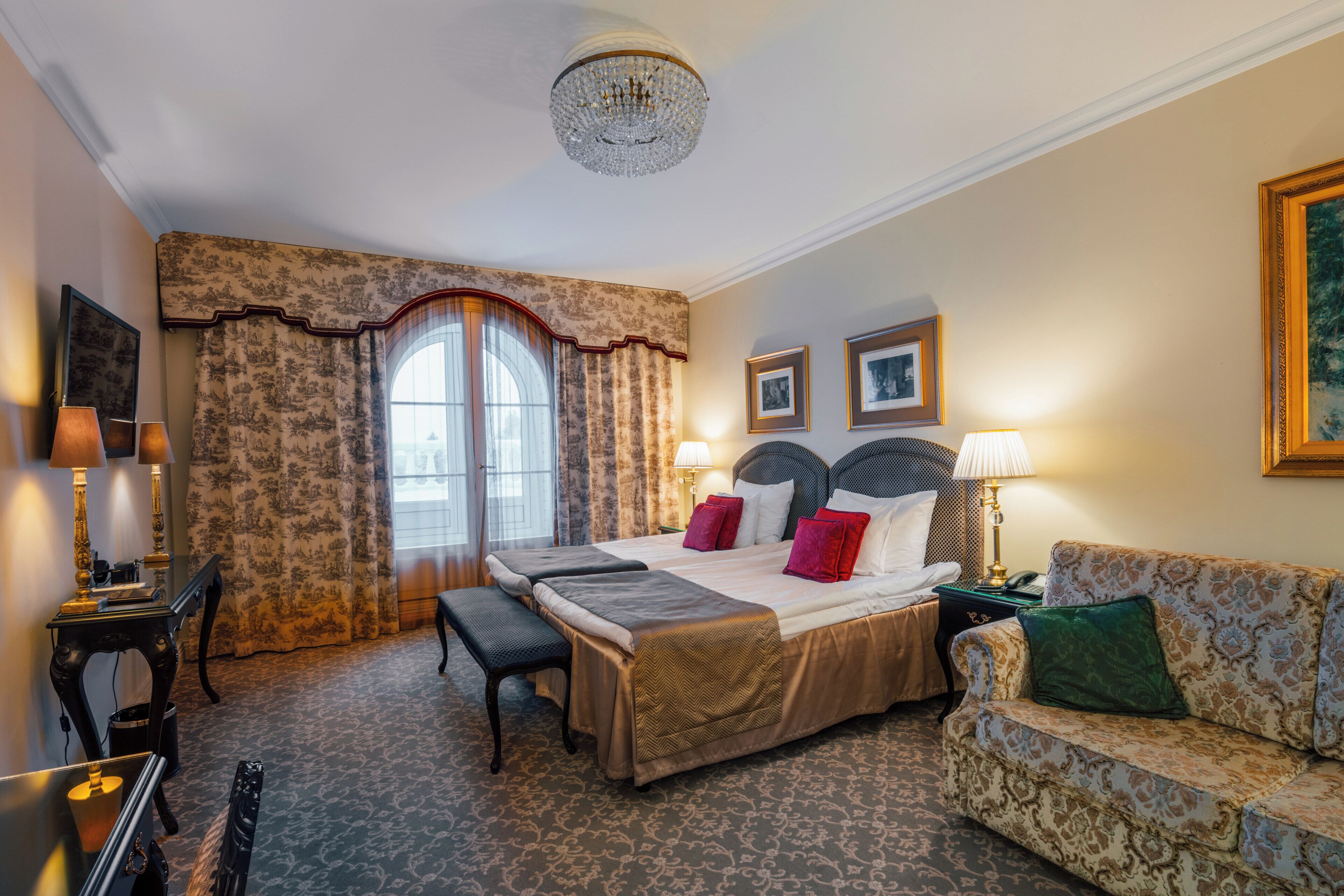 A hotel room with a large bed, a sofa, a desk, and a chandelier.