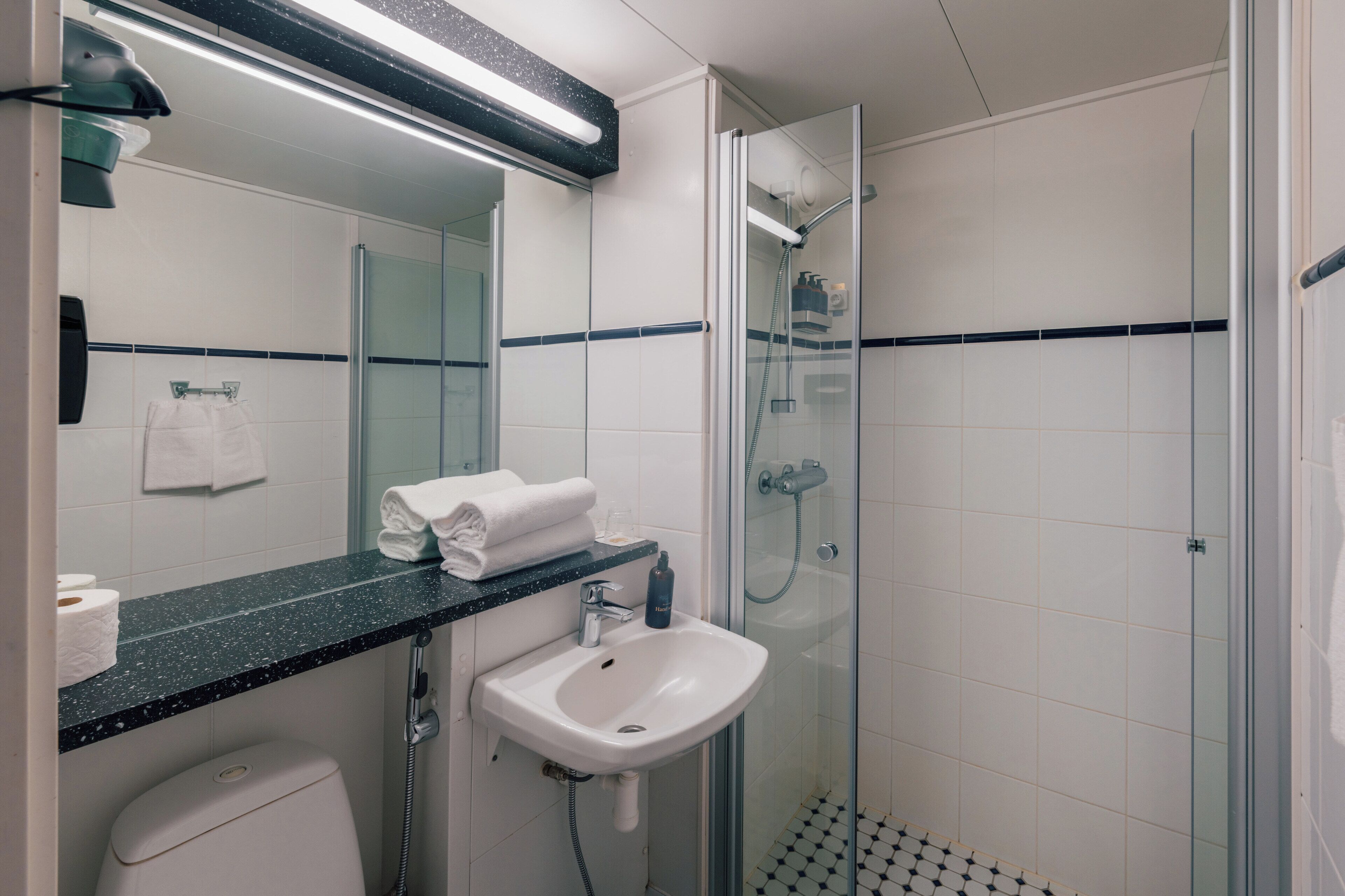Un baño moderno con ducha envuelta en vidrio, lavamanos blanco y encimera negra.