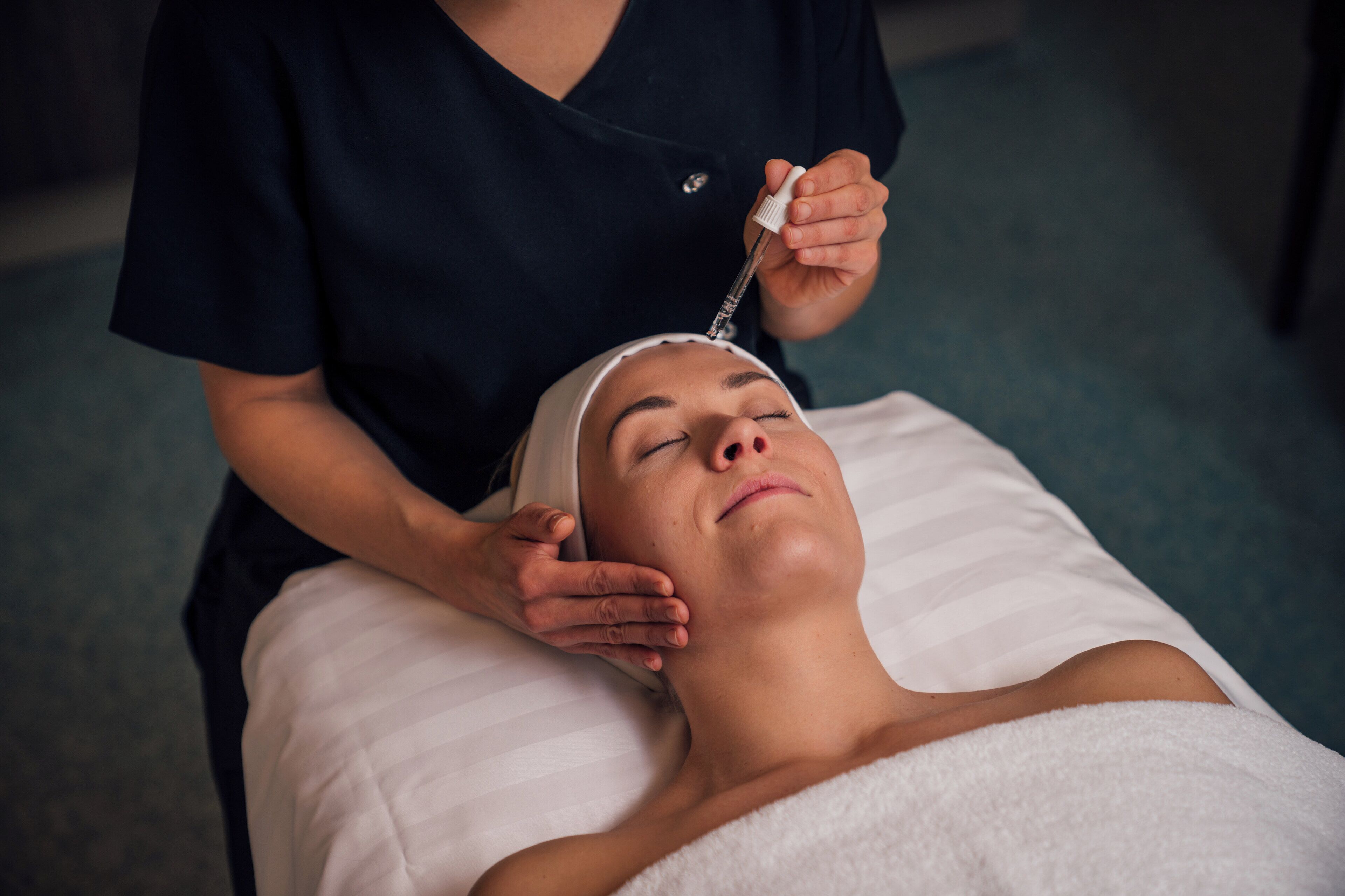 A person lying on a bed with a white towel, receiving a facial treatment from a professional in a dark uniform.