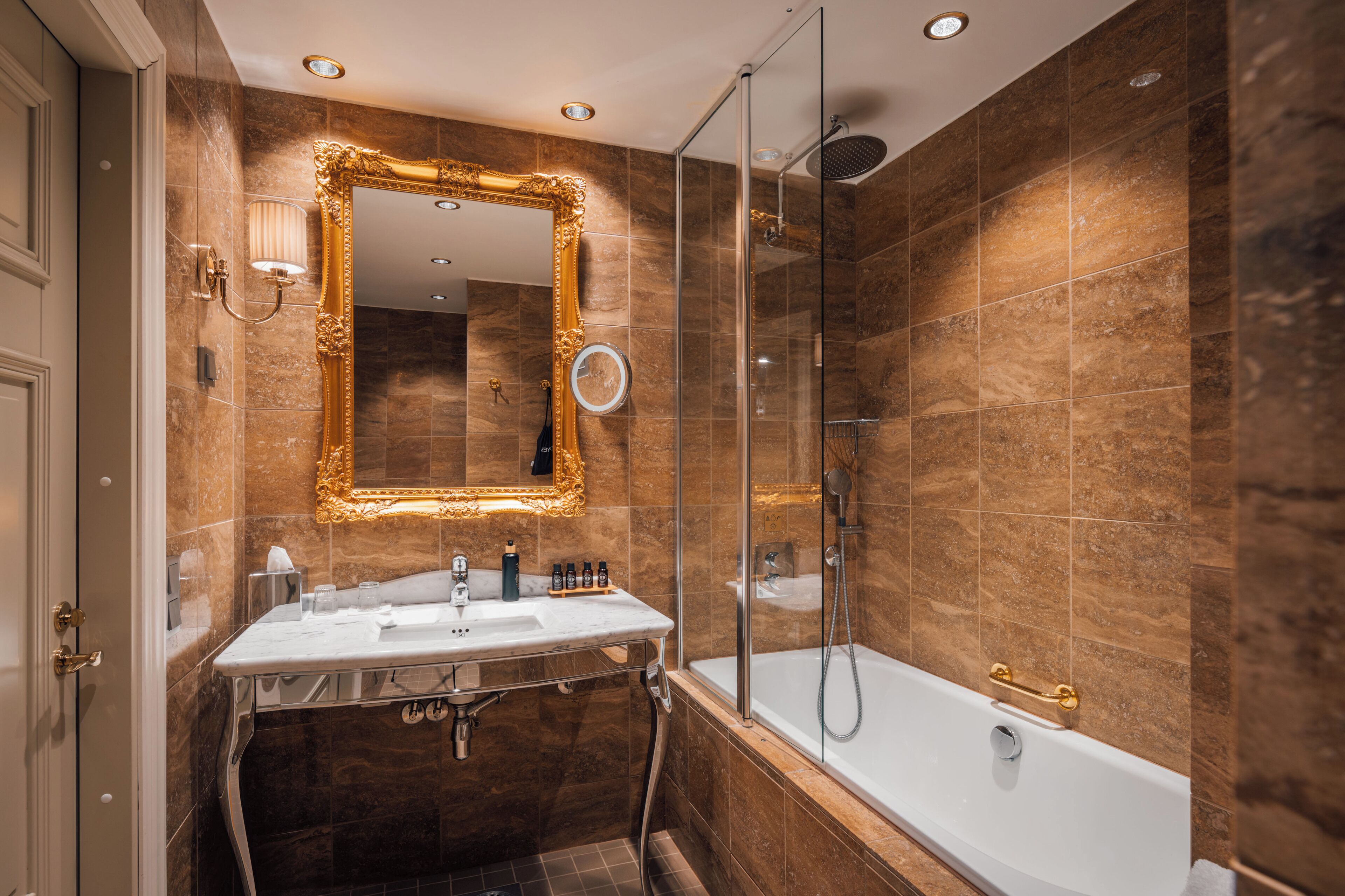 Une salle de bain avec un mur en marbre, un miroir encadré d’or, une baignoire blanche et une douche à parois de verre.