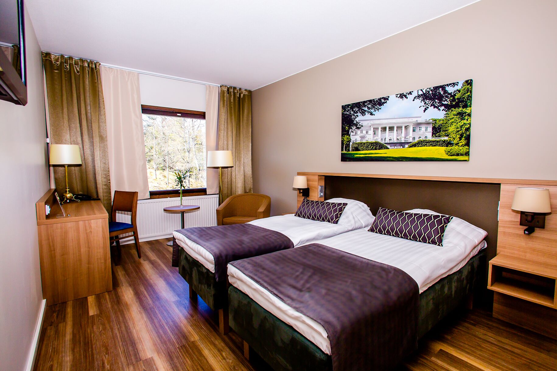 A hotel room with two beds, a desk, a chair, a window with curtains, and a painting on the wall.