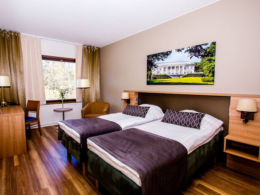 A hotel room with two beds, a desk, a chair, a window with curtains, and a painting on the wall.