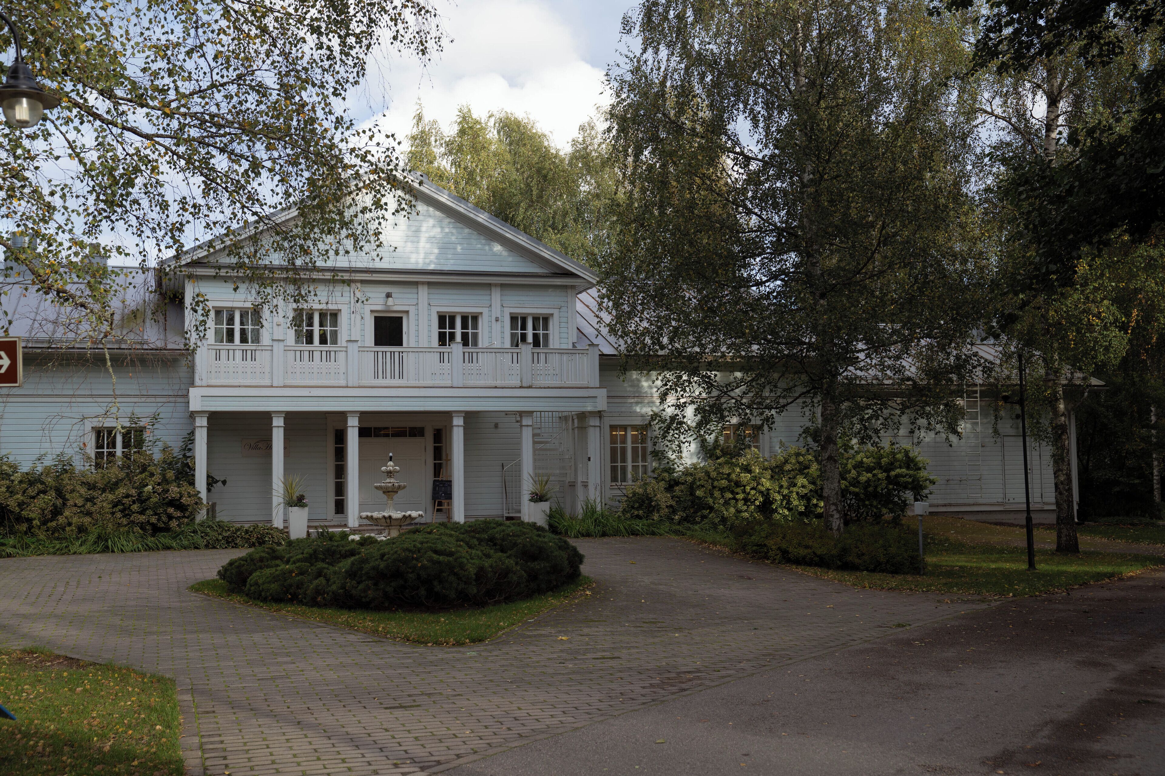 A white colonial-style building with a front porch, surrounded by trees and a paved driveway.