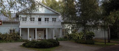 A white colonial-style building with a front porch, surrounded by trees and a paved driveway.