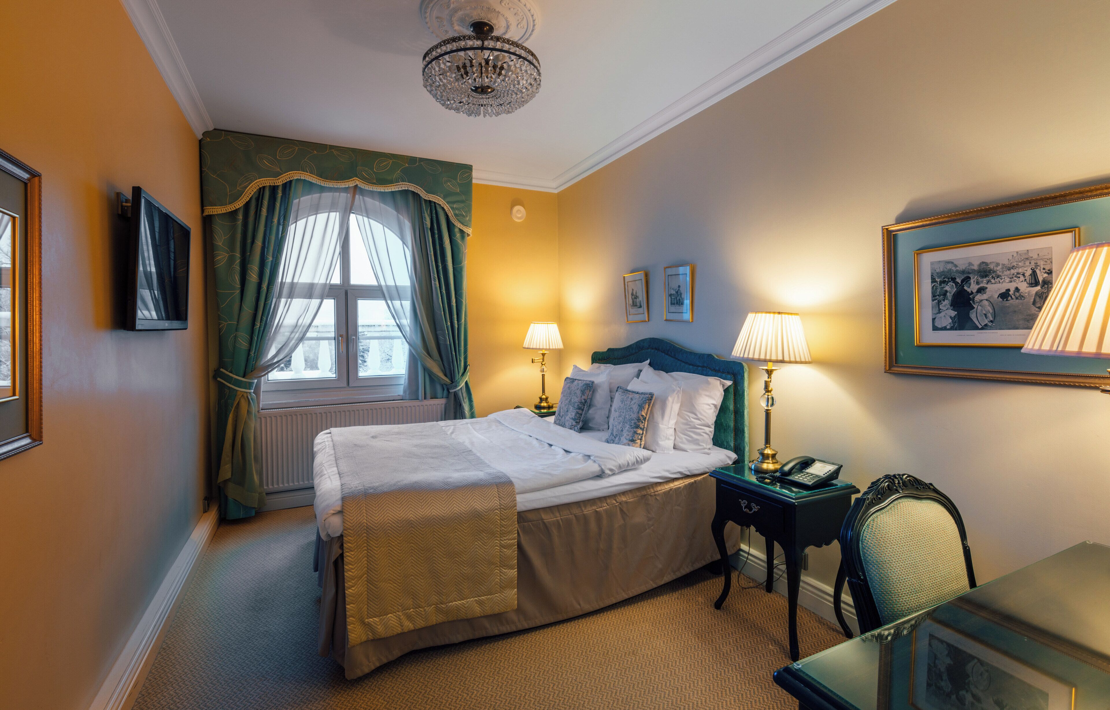 Een hotelkamer met een bed, twee bedlampen, een stoel, een tafel, een lijst aan de muur en een raam met gordijnen.