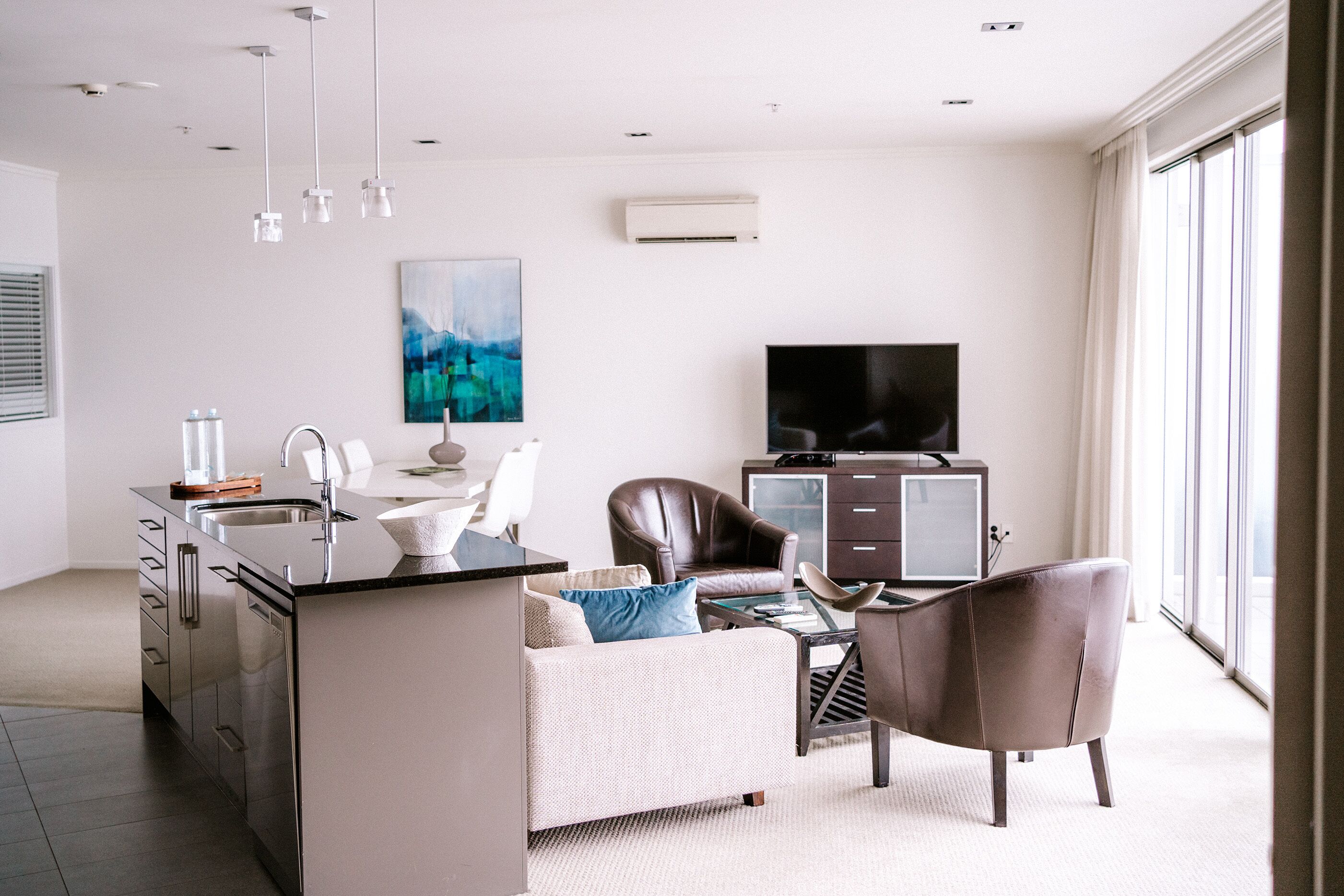 A modern living room with a kitchen area, a flat-screen TV, and a large window.