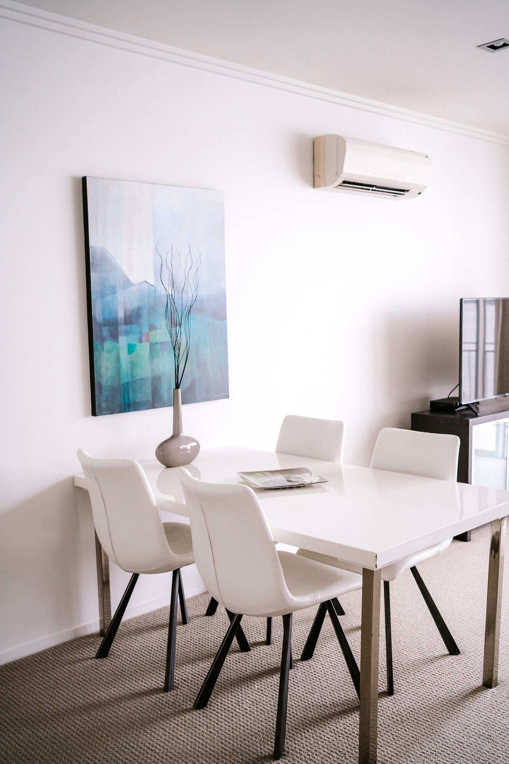 Een moderne eetruimte met een witte tafel en stoelen, een vaas met takken en een wandairconditioner.
