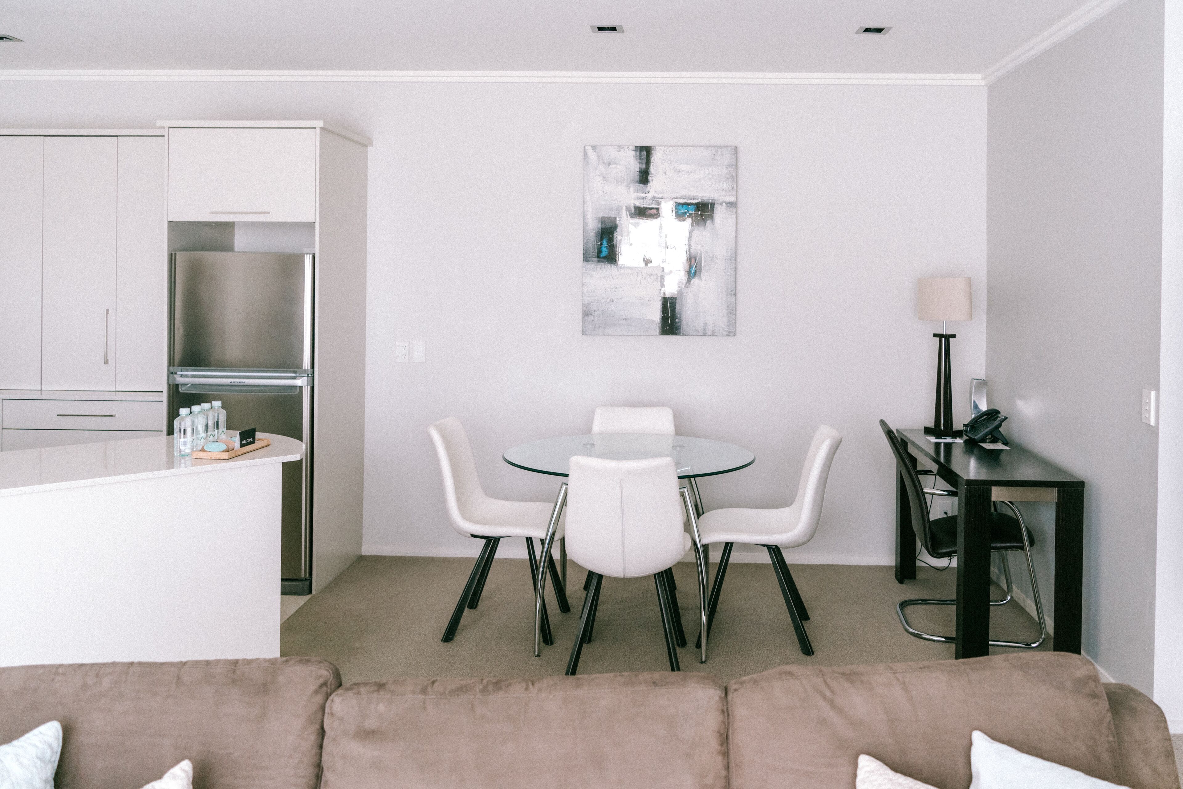 A modern kitchen with white cabinets and a stainless steel refrigerator, a dining area with a glass table and white chairs, and a small black side table with a lamp.