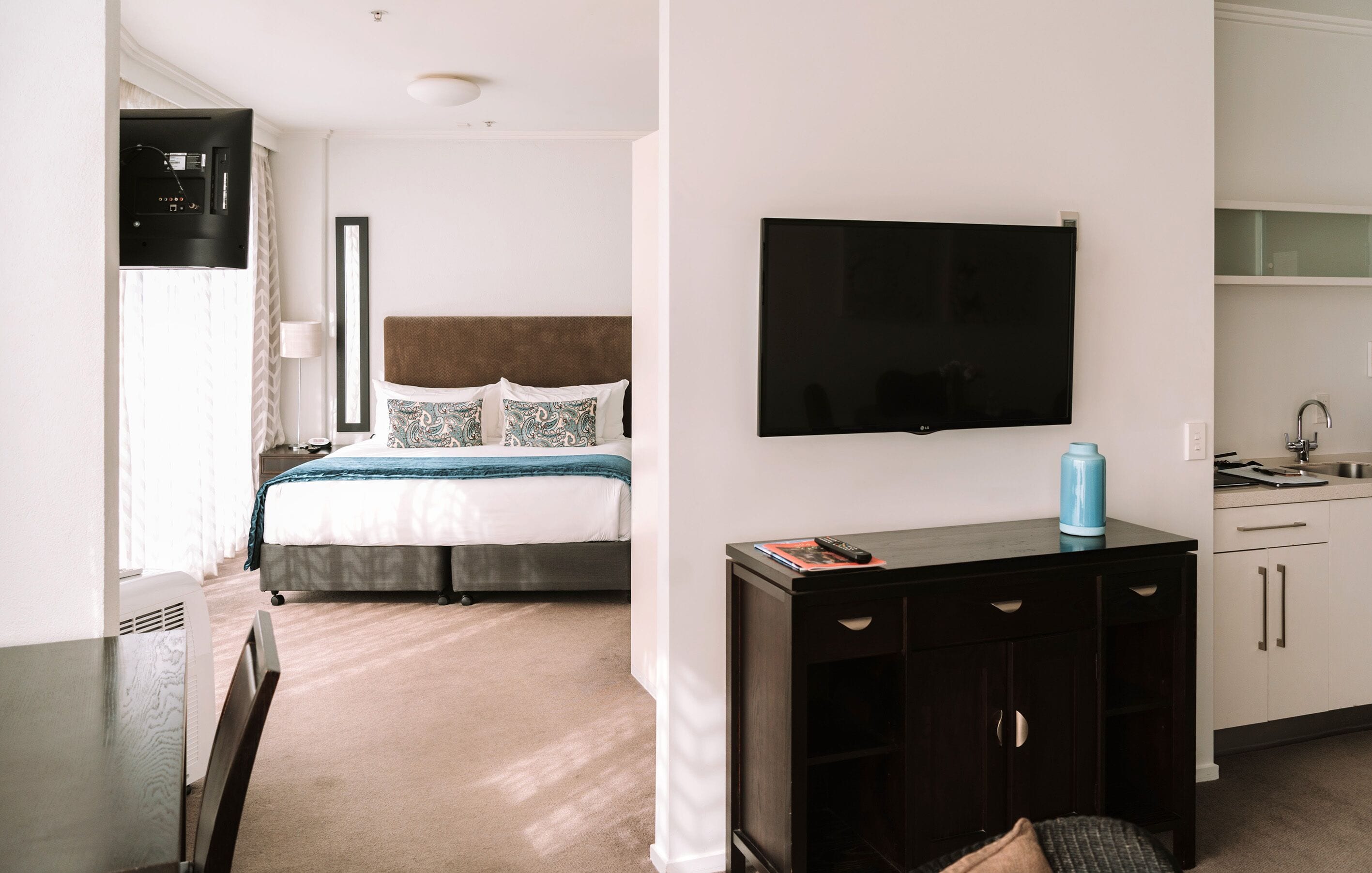 A hotel room with a bed, a TV mounted on the wall, a small table with a remote control, and a kitchenette area.